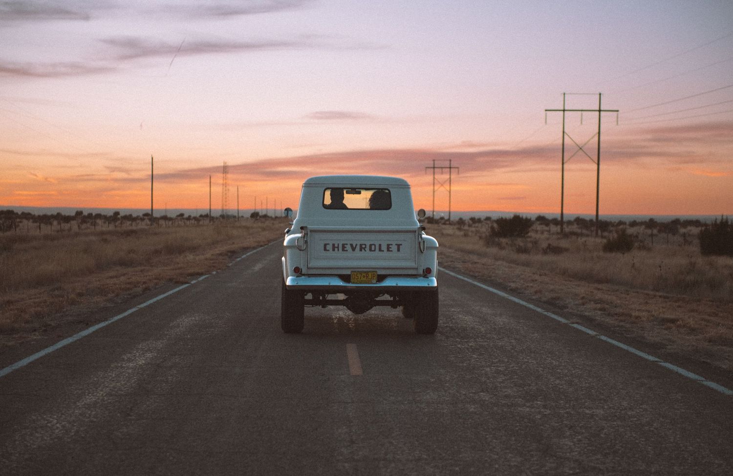 Vintage light blue Chevrolet pickup truck driving down an empty road at colorful sunset.