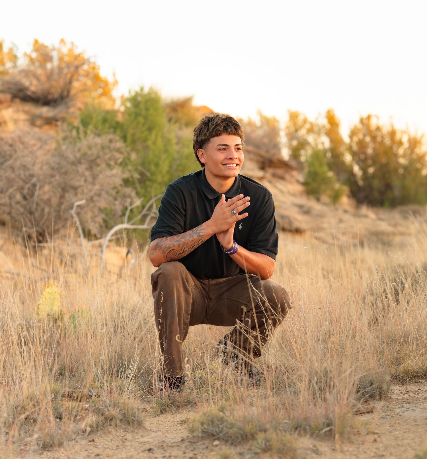 Young man crouching in golden desert field, smiling with tattooed arms, wearing black polo and brown pants.