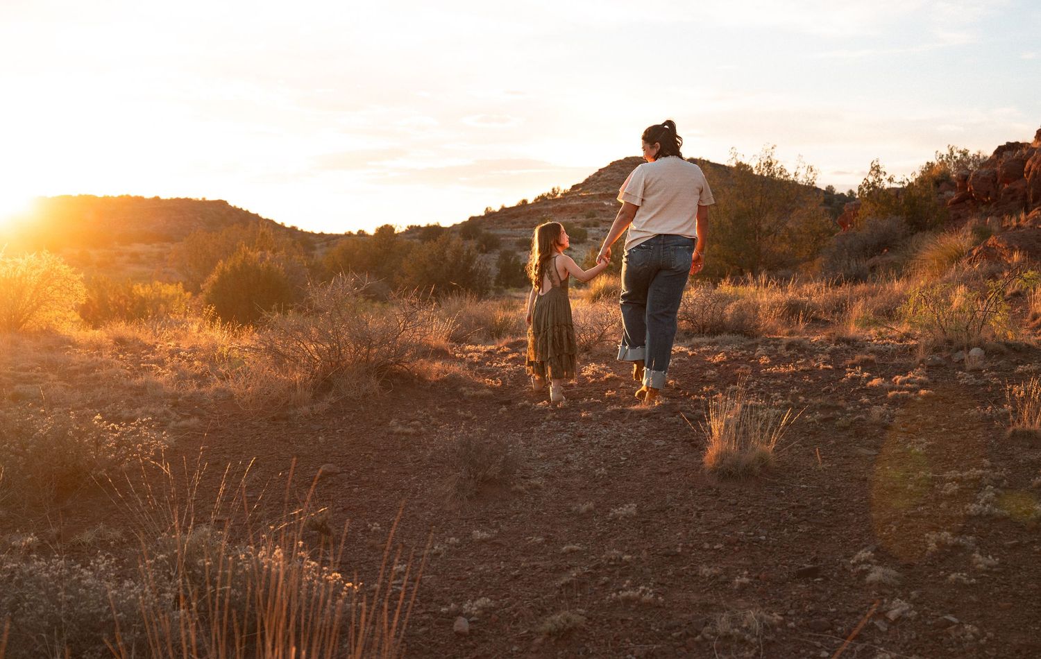 Adult and child walking hand in hand on a dirt trail at sunset in a desert landscape.