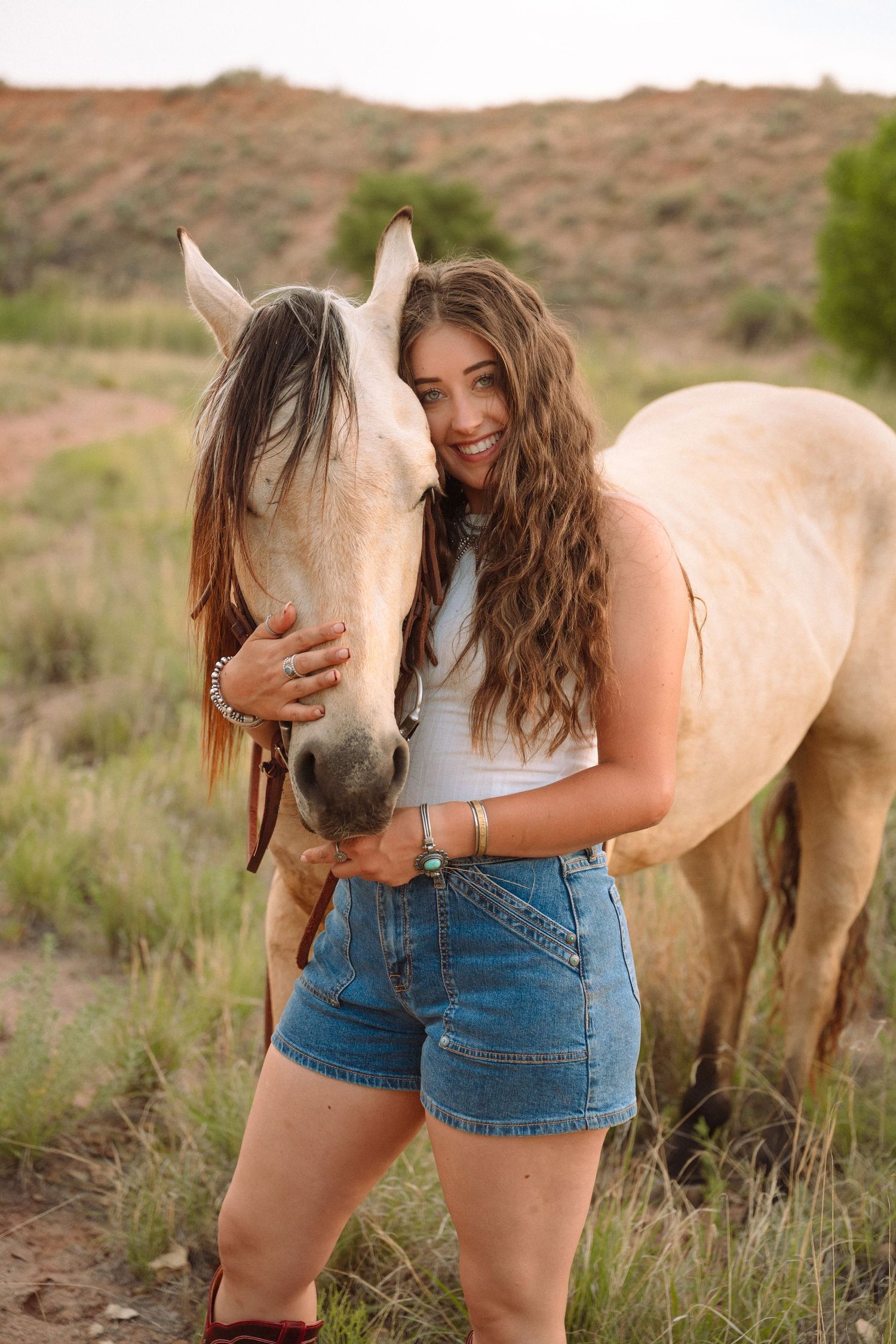 Smiling young woman in denim shorts hugging a palomino horse in a scenic desert field.