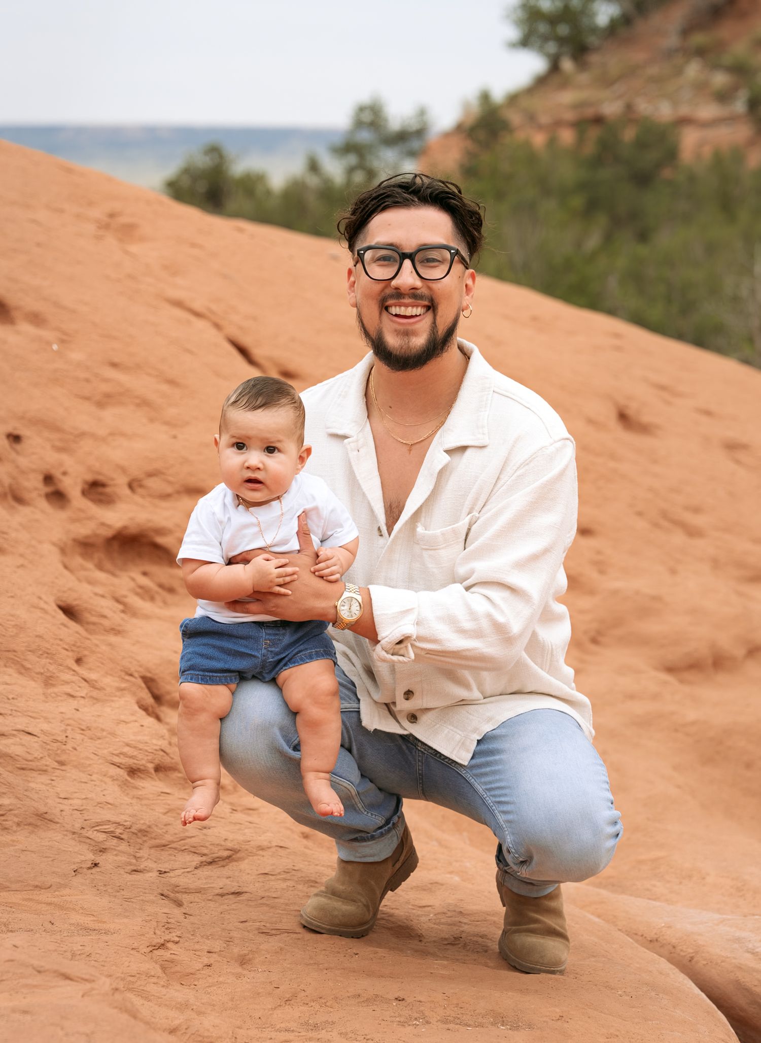 Smiling man in white shirt crouching with baby on red sandstone rock outdoors.