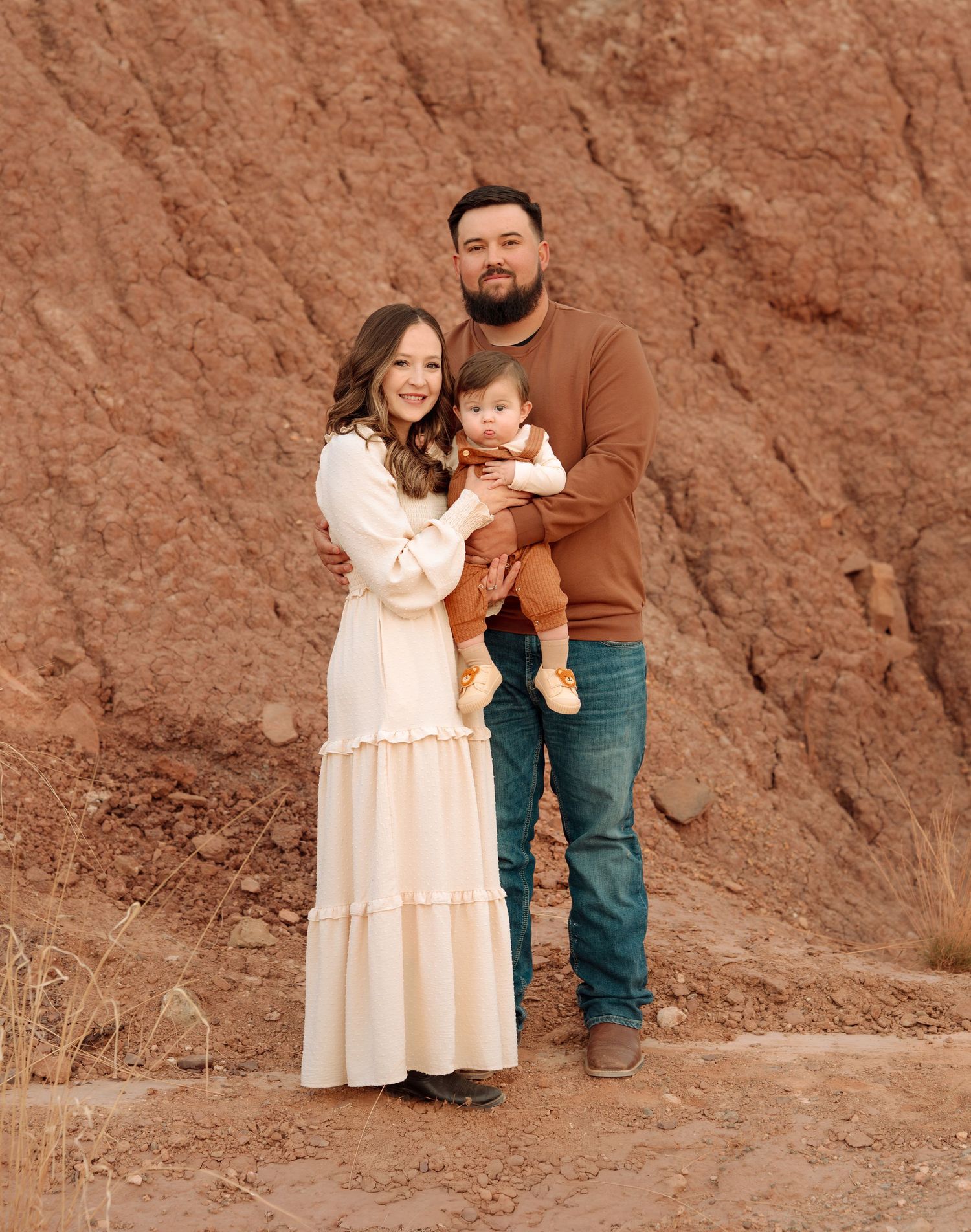 A smiling couple holds their baby in front of red rock cliffs in a warm family portrait.