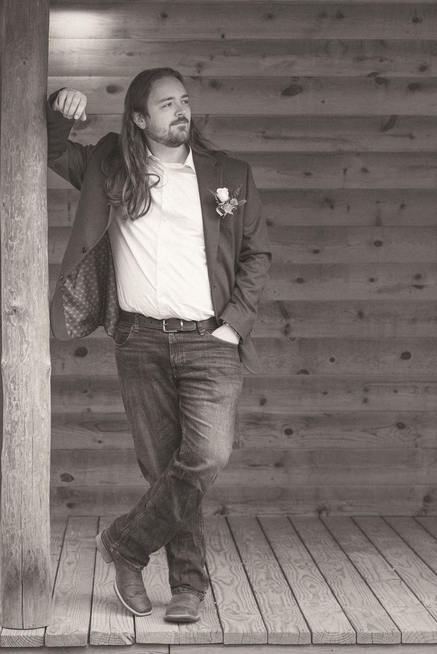 Groom in blazer and jeans leaning against log cabin wall in black and white wedding photo.