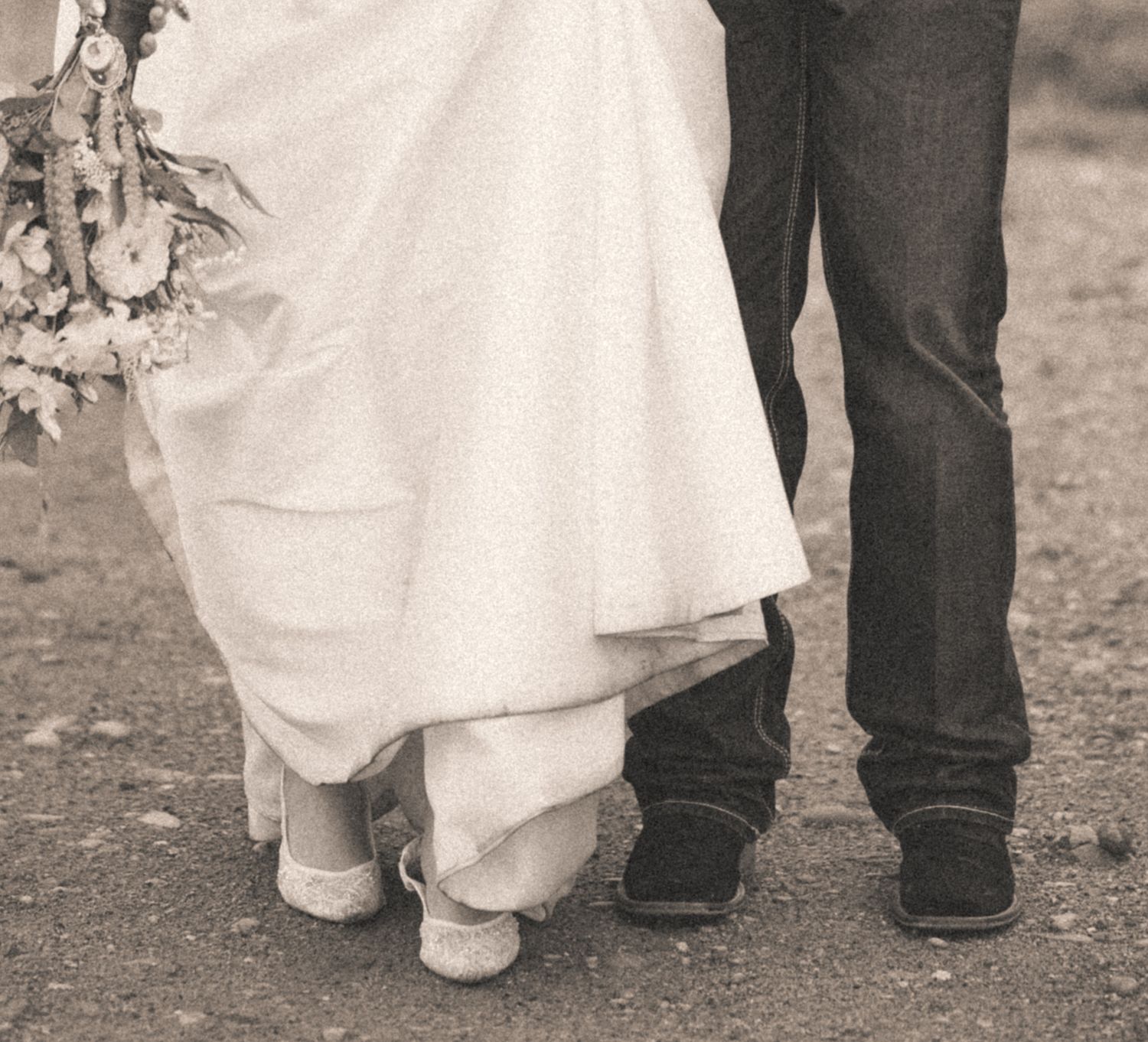 Bride and groom walking together, bride in white gown holding bouquet, groom in jeans and boots, black and white photo.