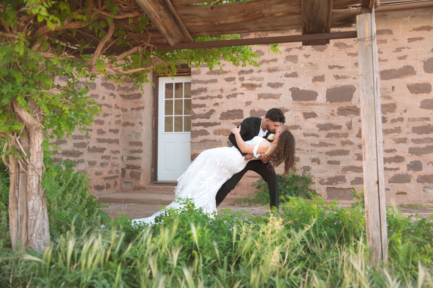 Groom dips bride dramatically in a lush green garden outside a stone venue.