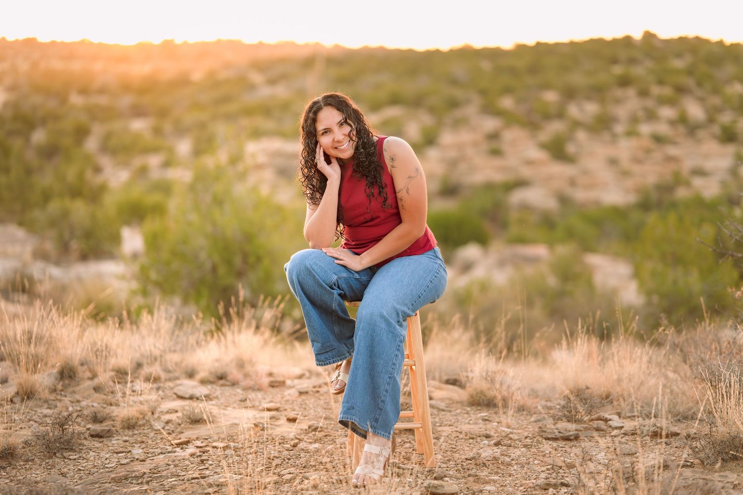 Woman in red top and jeans sits on stool in golden desert landscape at sunset.