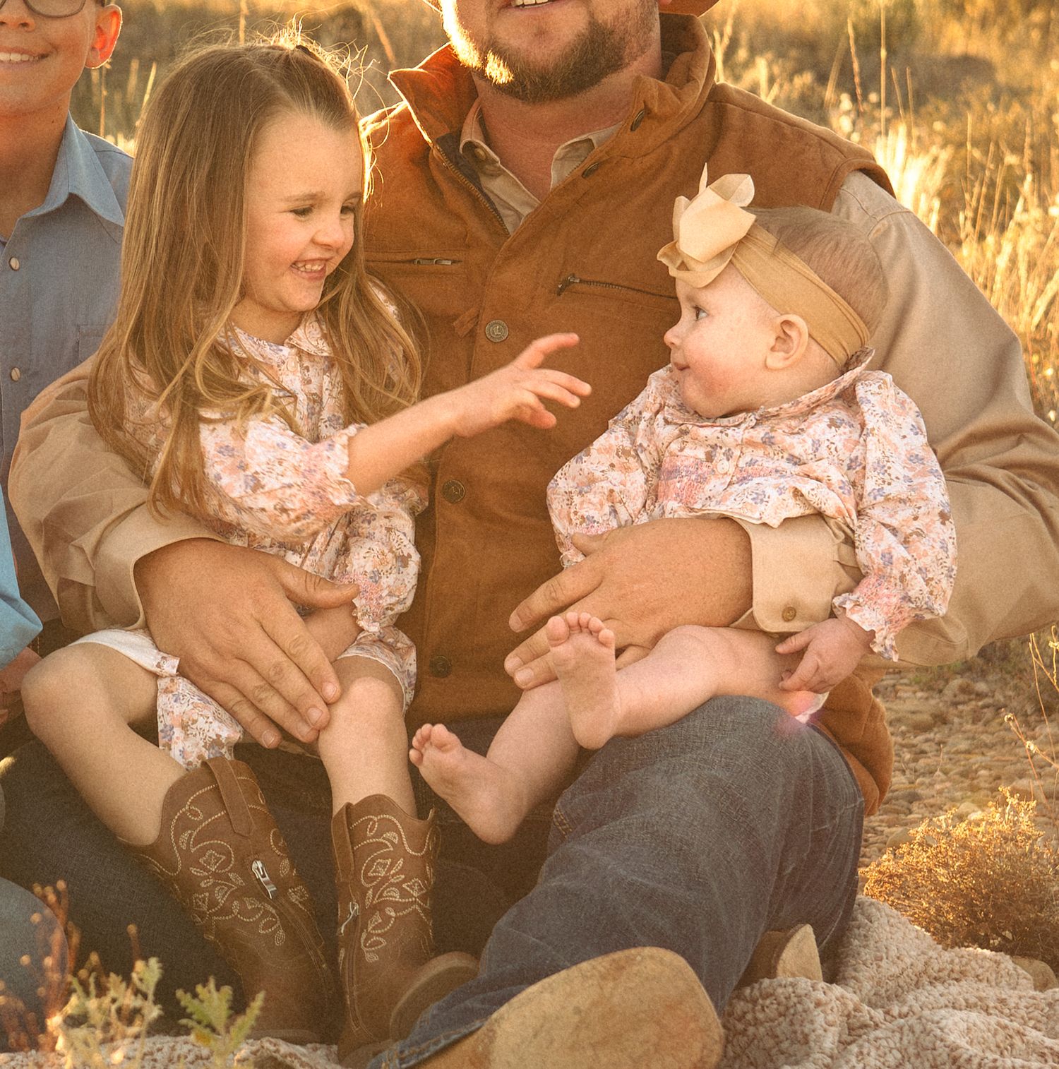 Young girl interacting with baby sibling on fathers lap in golden field at sunset.