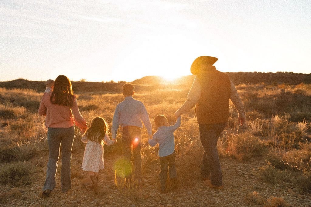 Silhouette of family of five holding hands walking toward golden sunset in open desert field.