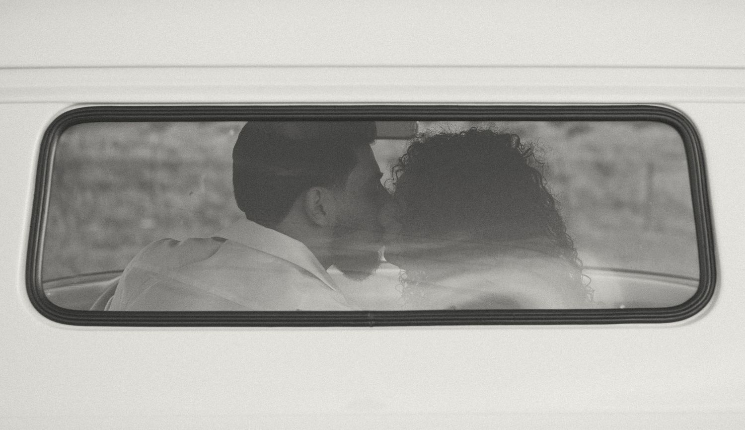 Black and white romantic photo of couple kissing viewed through rear van window.