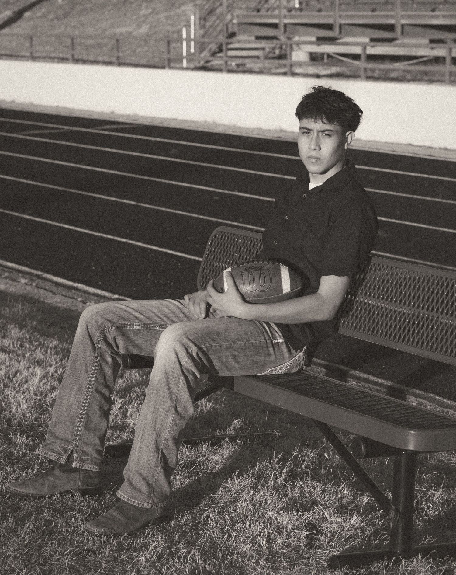 Black and white senior portrait of young man holding football on bench near stadium track.