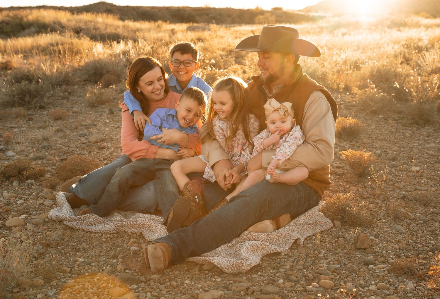 Happy family of six sitting together on a blanket in a golden sunlit desert field.