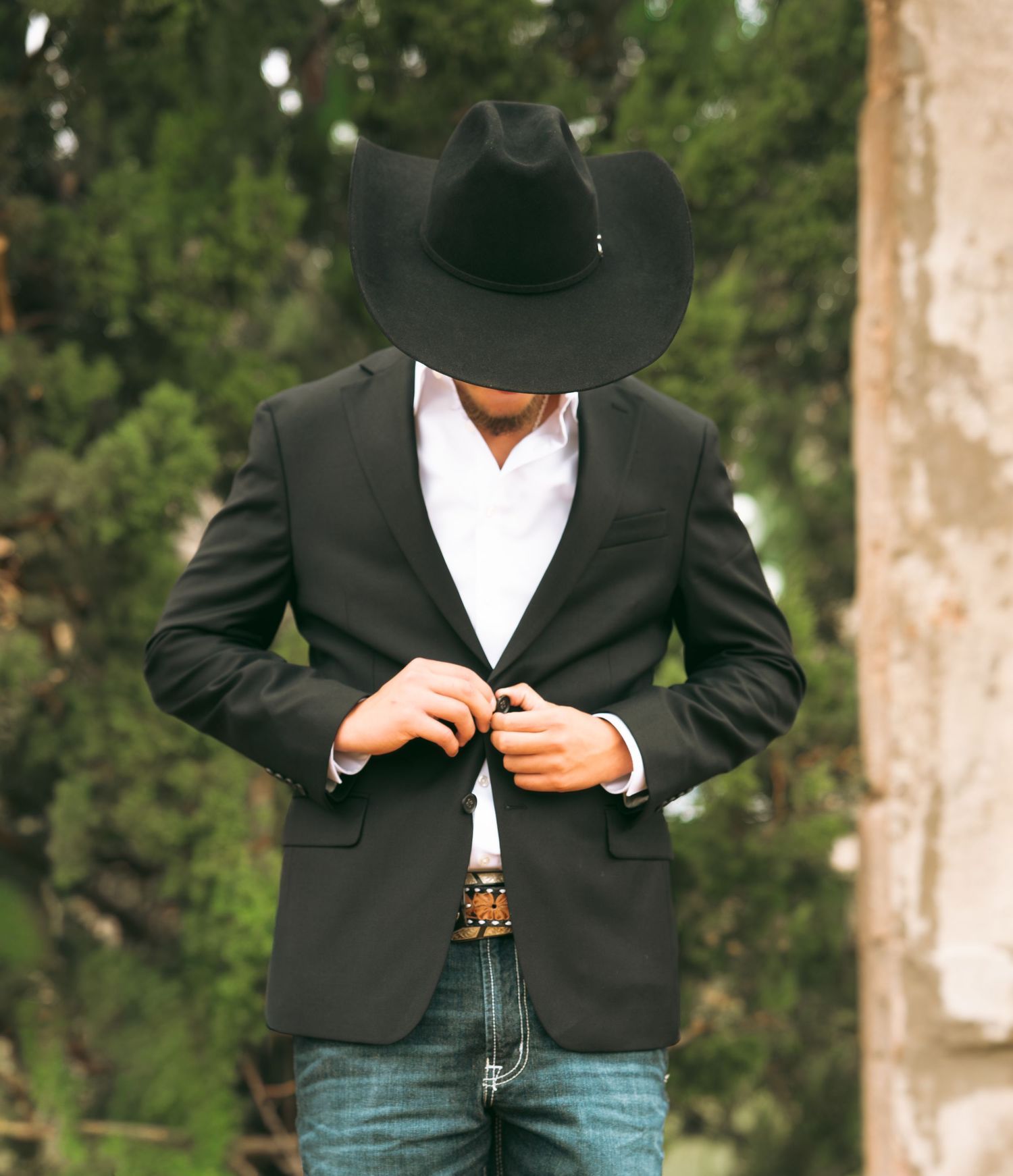 Man in black cowboy hat and blazer buttoning jacket outdoors with trees in background.
