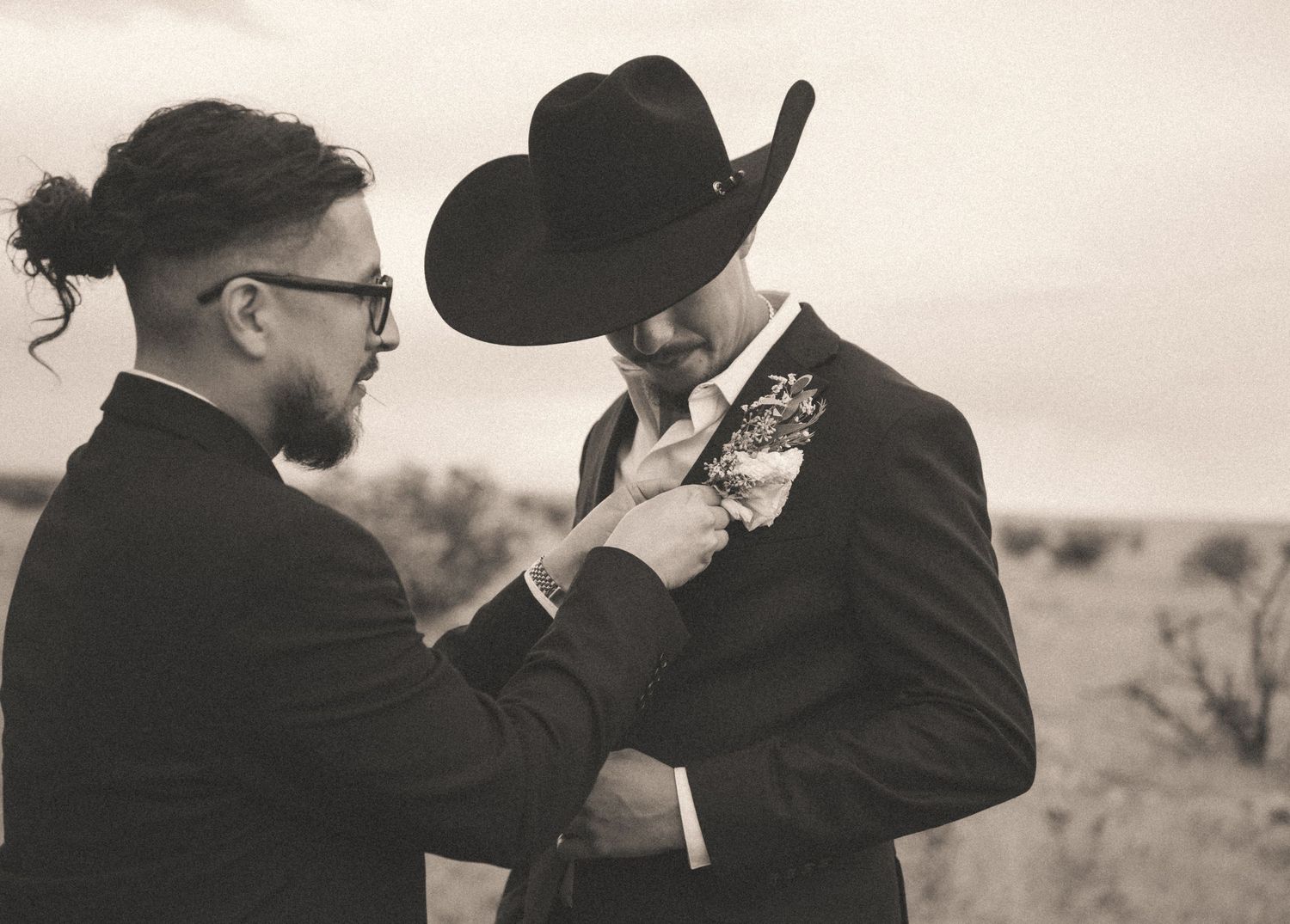 Black and white photo of groomsman pinning boutonniere on groom wearing black cowboy hat and suit.