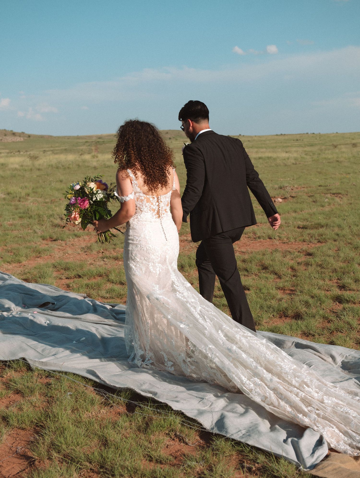 Bride and groom walk hand in hand across a scenic open green meadow.