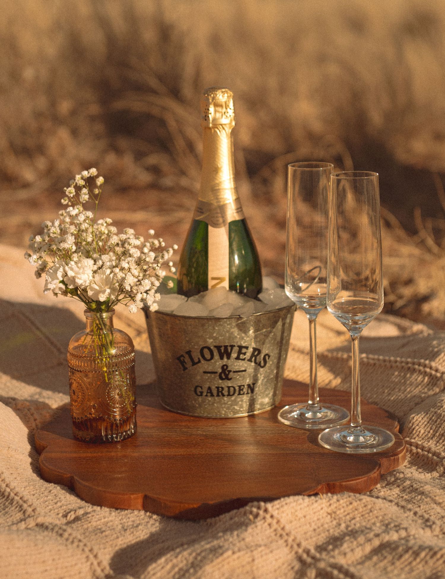 Champagne bottle in ice bucket with flutes and baby's breath flowers on wooden tray outdoors.