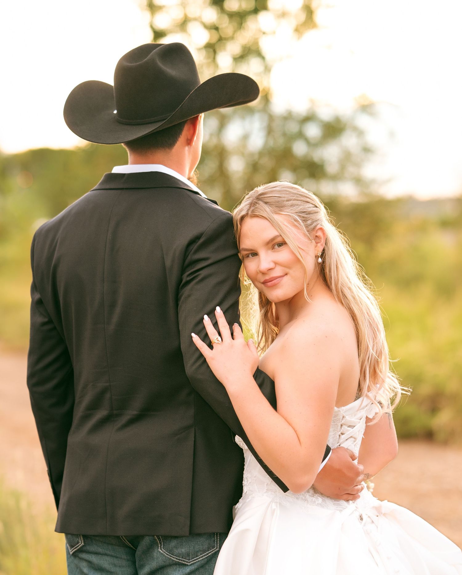 Smiling bride in lace gown embracing groom in black cowboy hat on rustic outdoor path.