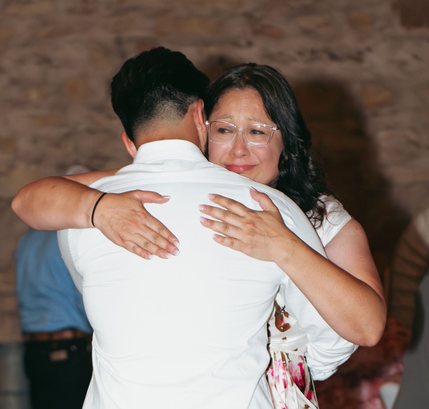 A woman with glasses warmly hugs a man in a white shirt at a wedding reception.