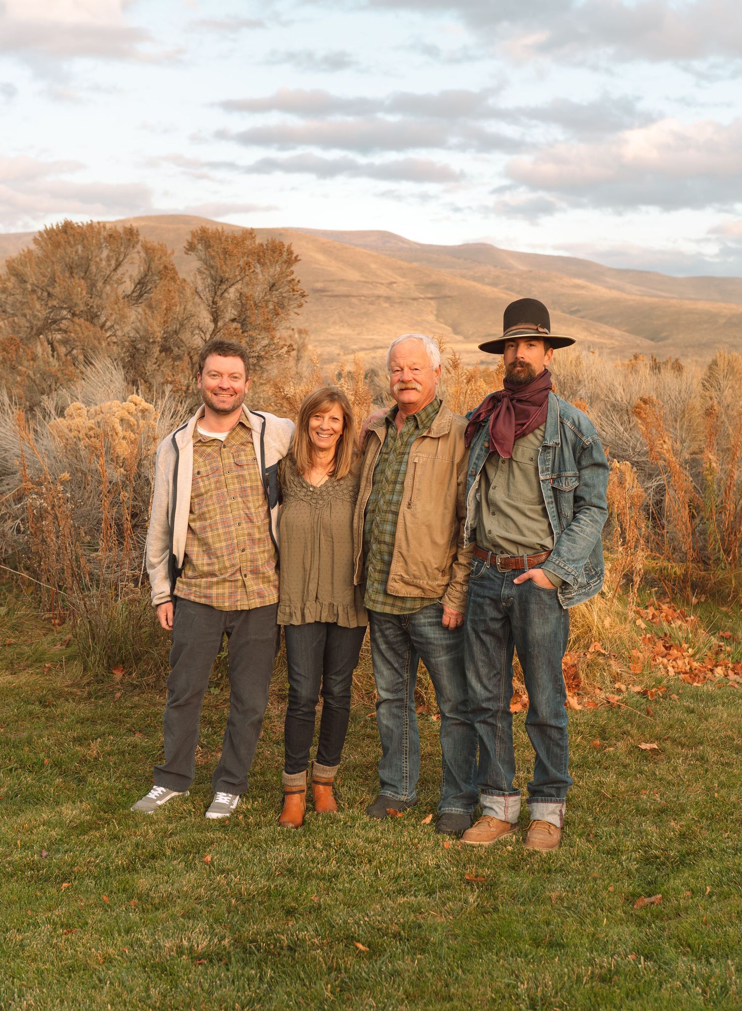 Group of four adults posing together on green lawn with rolling hills and autumn foliage in background.