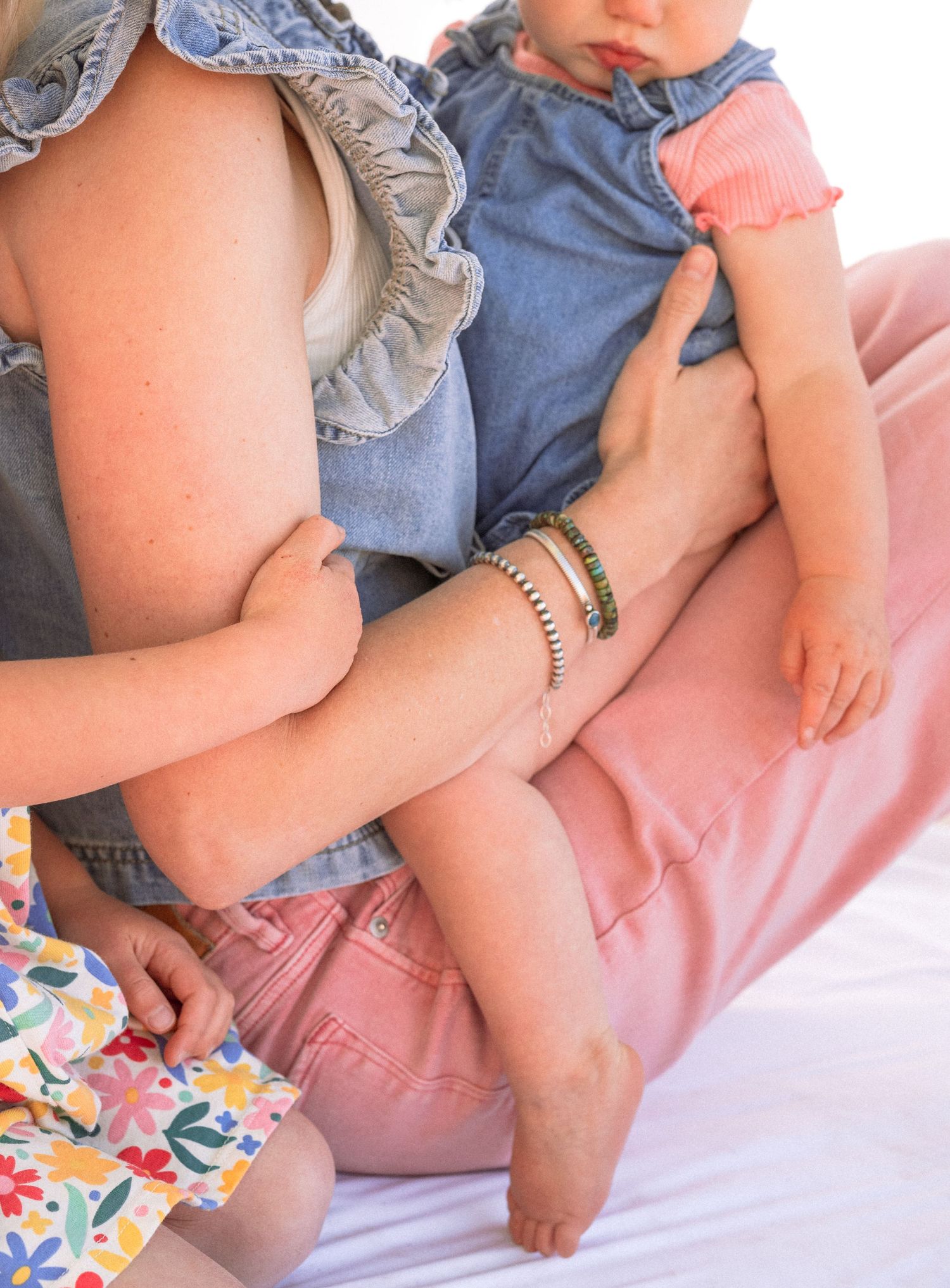 A mother holds a baby in a denim romper while a toddler in a floral dress sits nearby, all in soft pink tones.