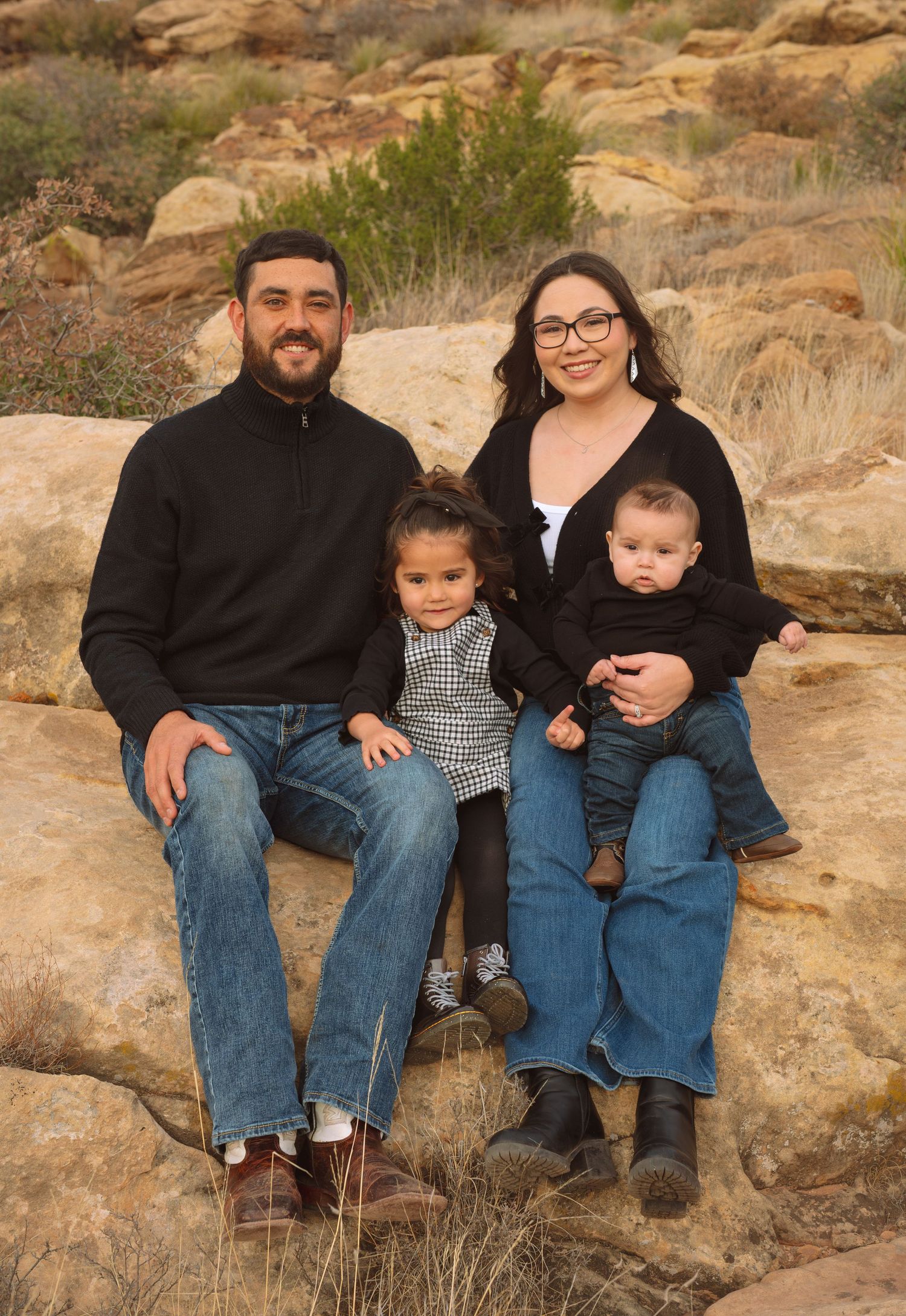 Family of four with toddler and baby posing on rocks in desert landscape for outdoor family portrait.