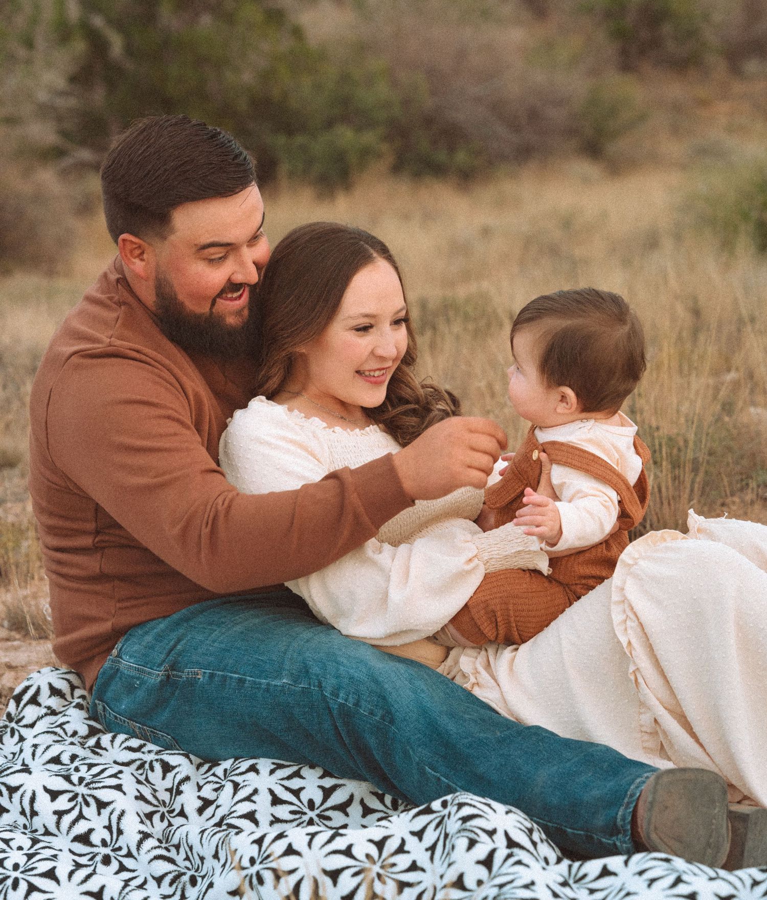 Happy young family of three sitting on floral blanket outdoors with parents smiling at baby.