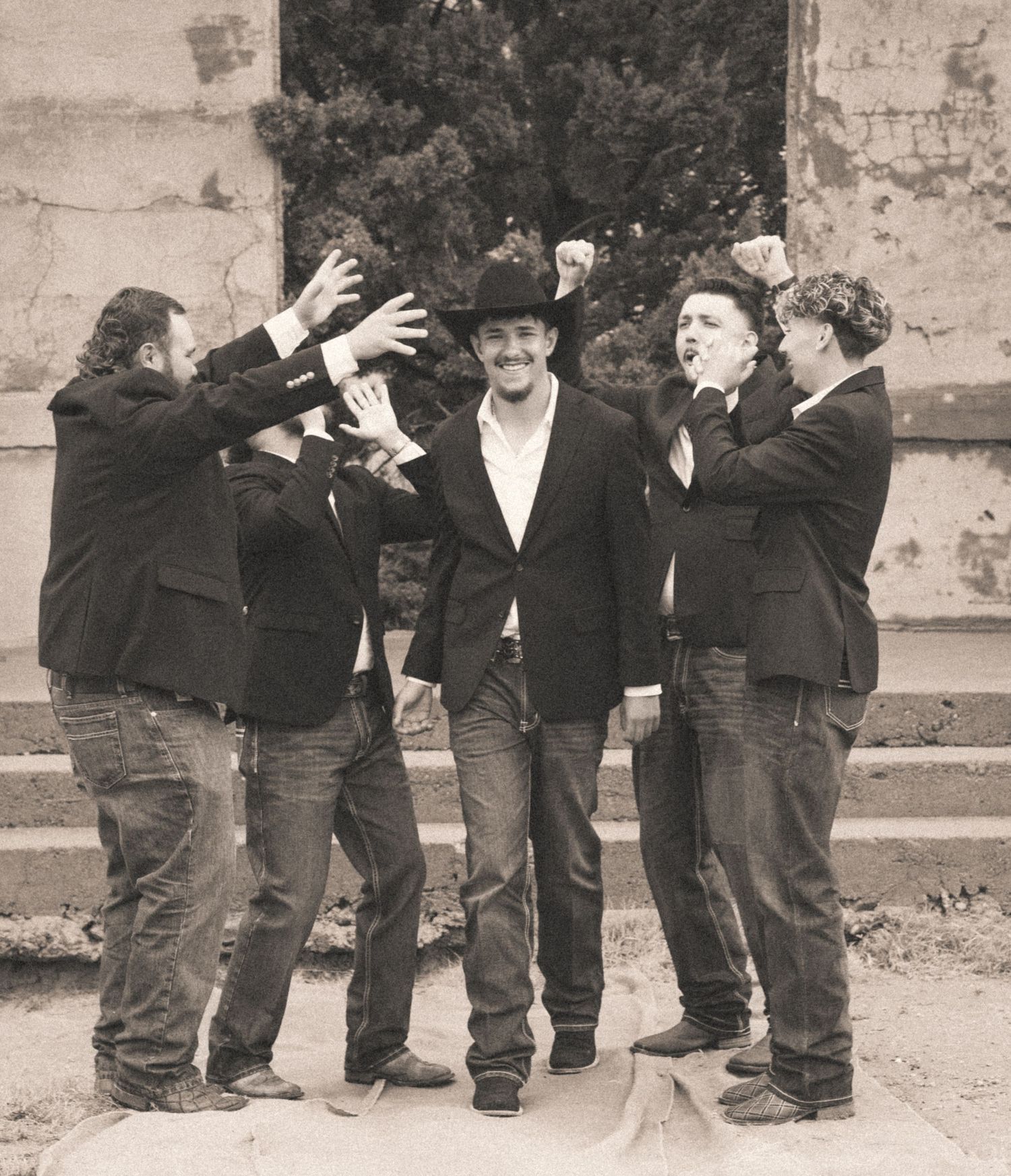 Groom in black cowboy hat celebrated by groomsmen in black suits in a fun wedding party photo.