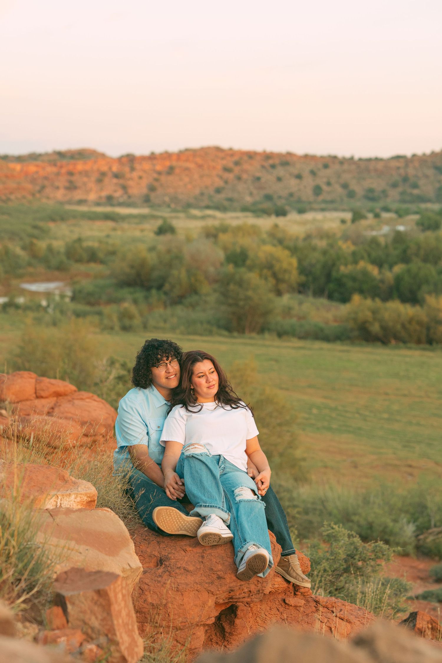 Couple sitting together on red rocks enjoying a scenic sunset view over a lush green valley landscape.