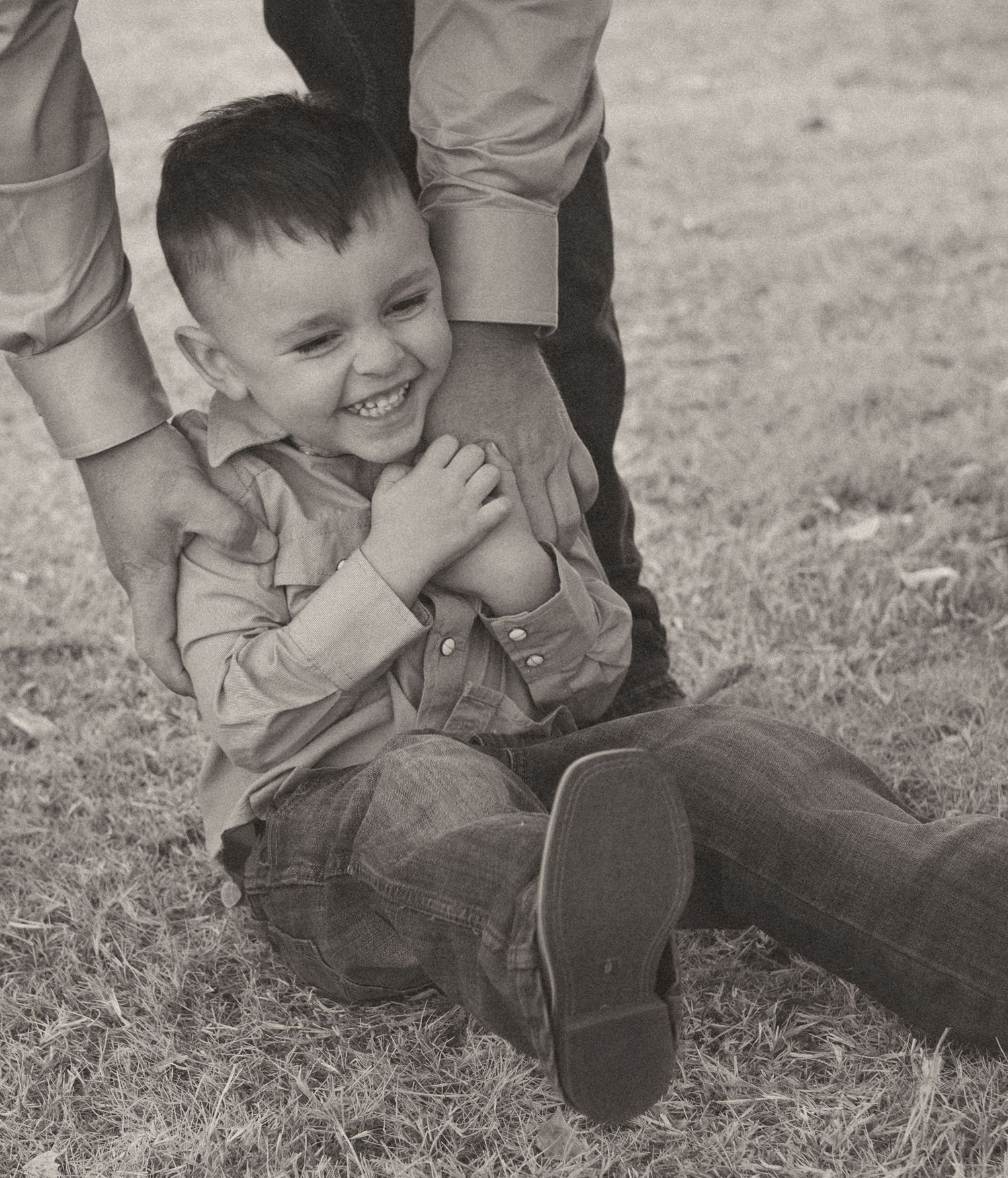 A joyful young boy laughs while being playfully held on grass in a black and white photo.