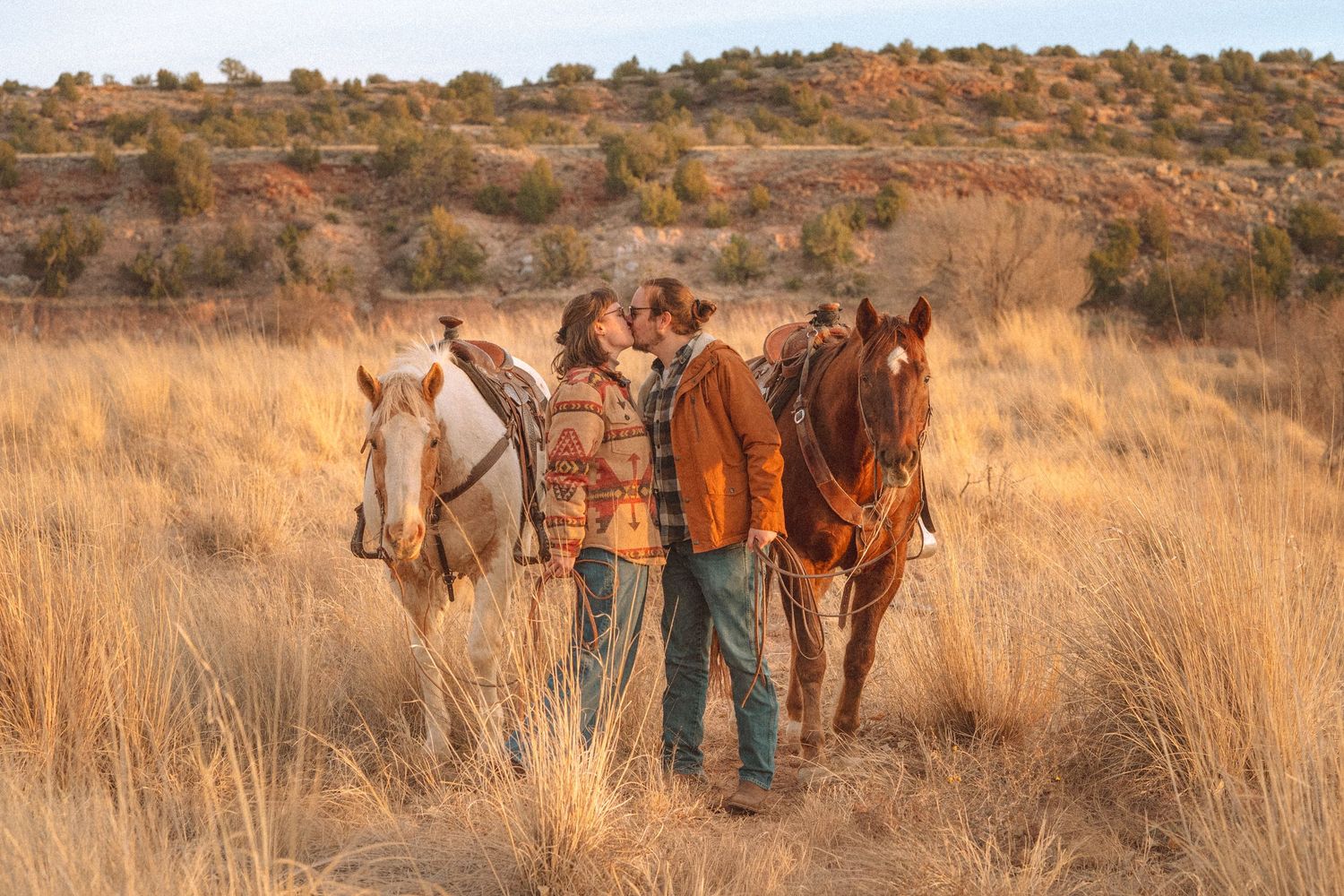 Couple kissing beside horses in golden prairie grass.