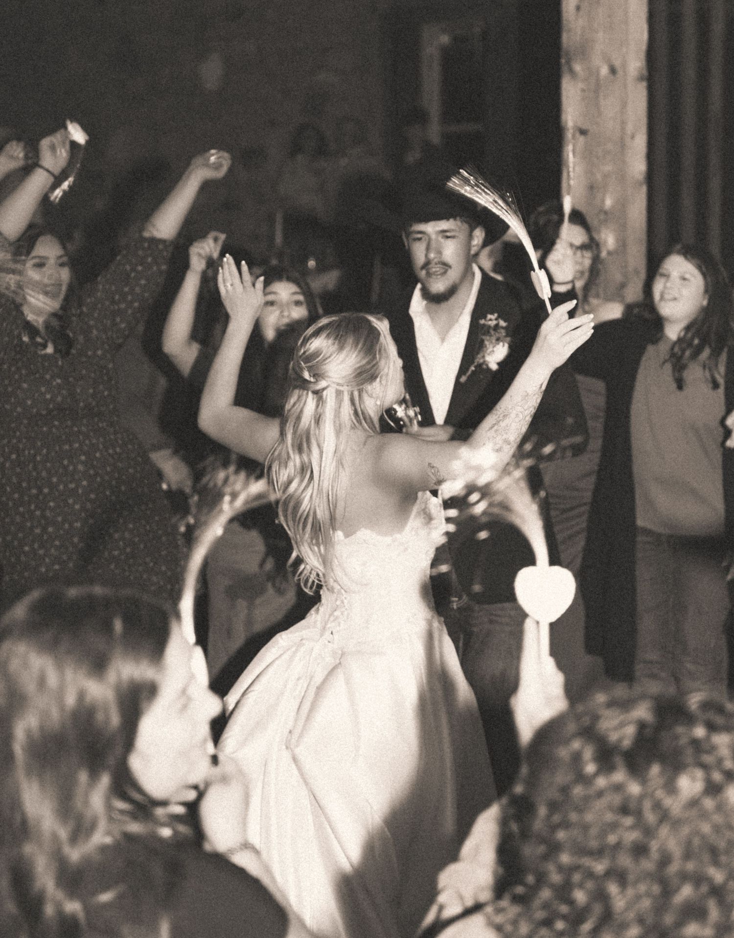 Bride and groom dancing at rustic wedding reception surrounded by cheering guests in sepia tone.