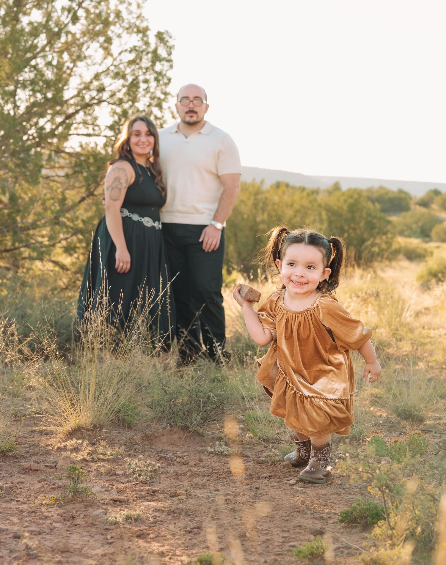 Parents stand in sunlit field watching their young daughter in golden dress run toward camera outdoors.