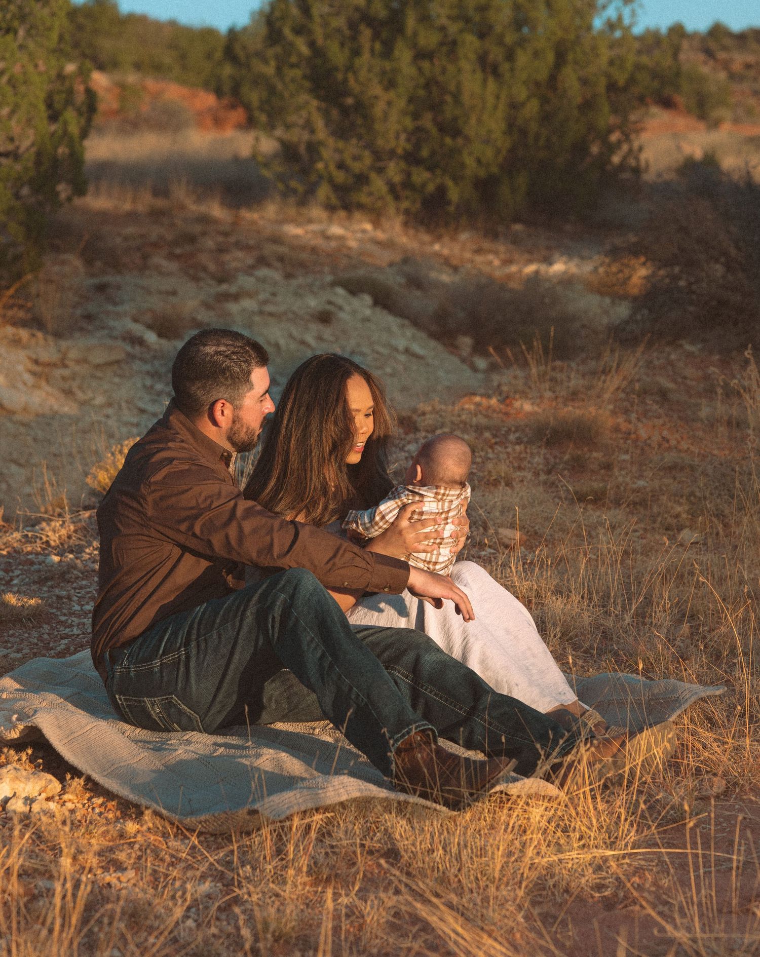 Young couple sitting on blanket in golden field holding newborn baby during sunset outdoor family photo session.