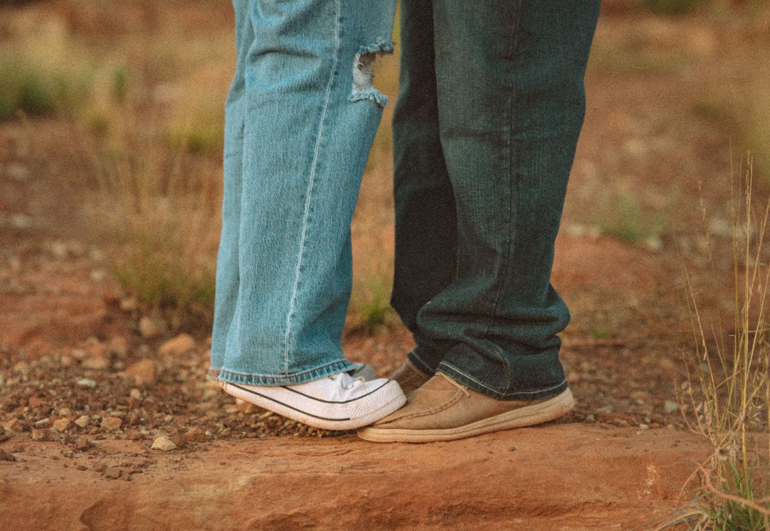 Two people standing close together on red dirt, wearing jeans and casual shoes in a desert landscape.