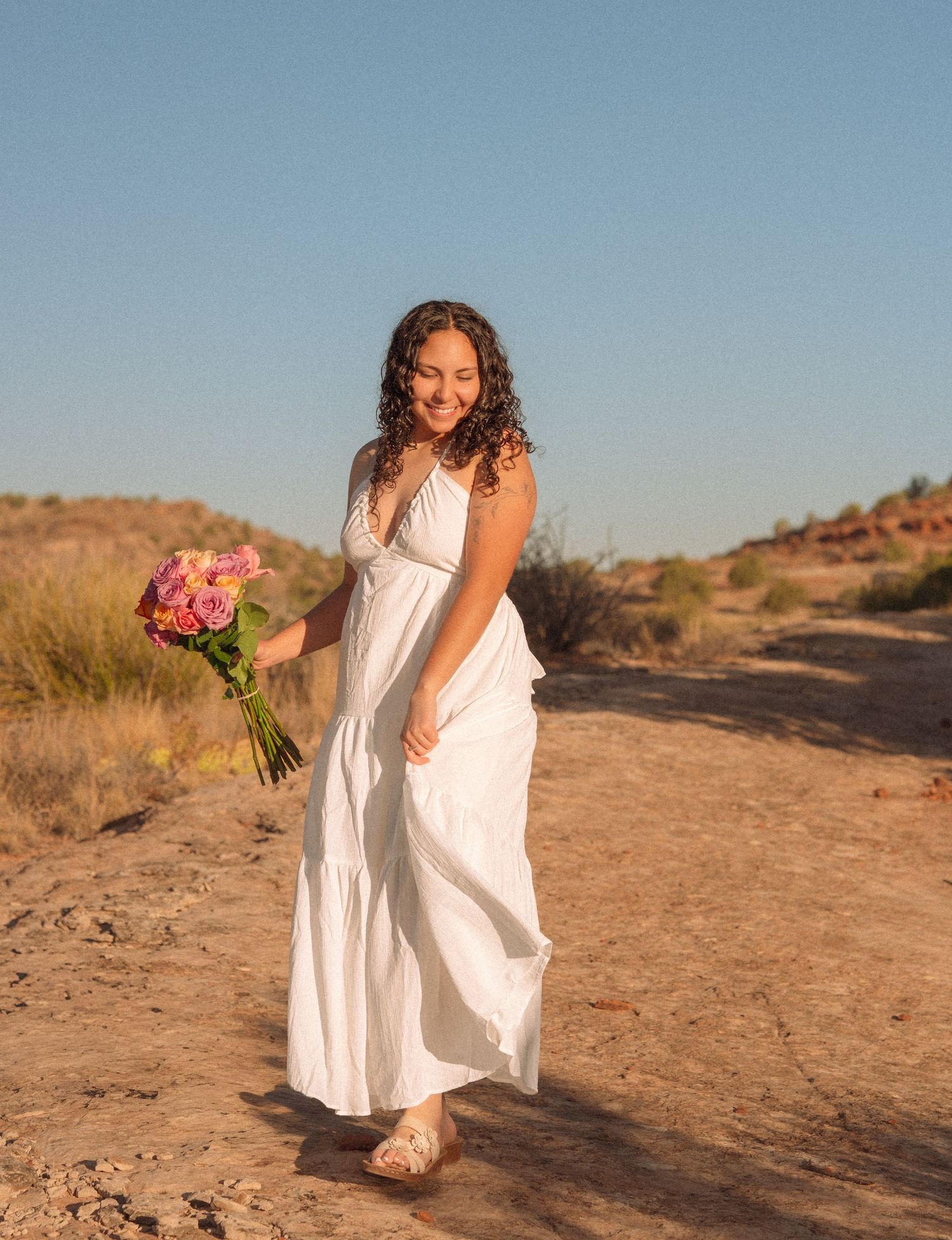 Woman in white maxi dress holding pink bouquet smiling outdoors in desert landscape on sunny day.