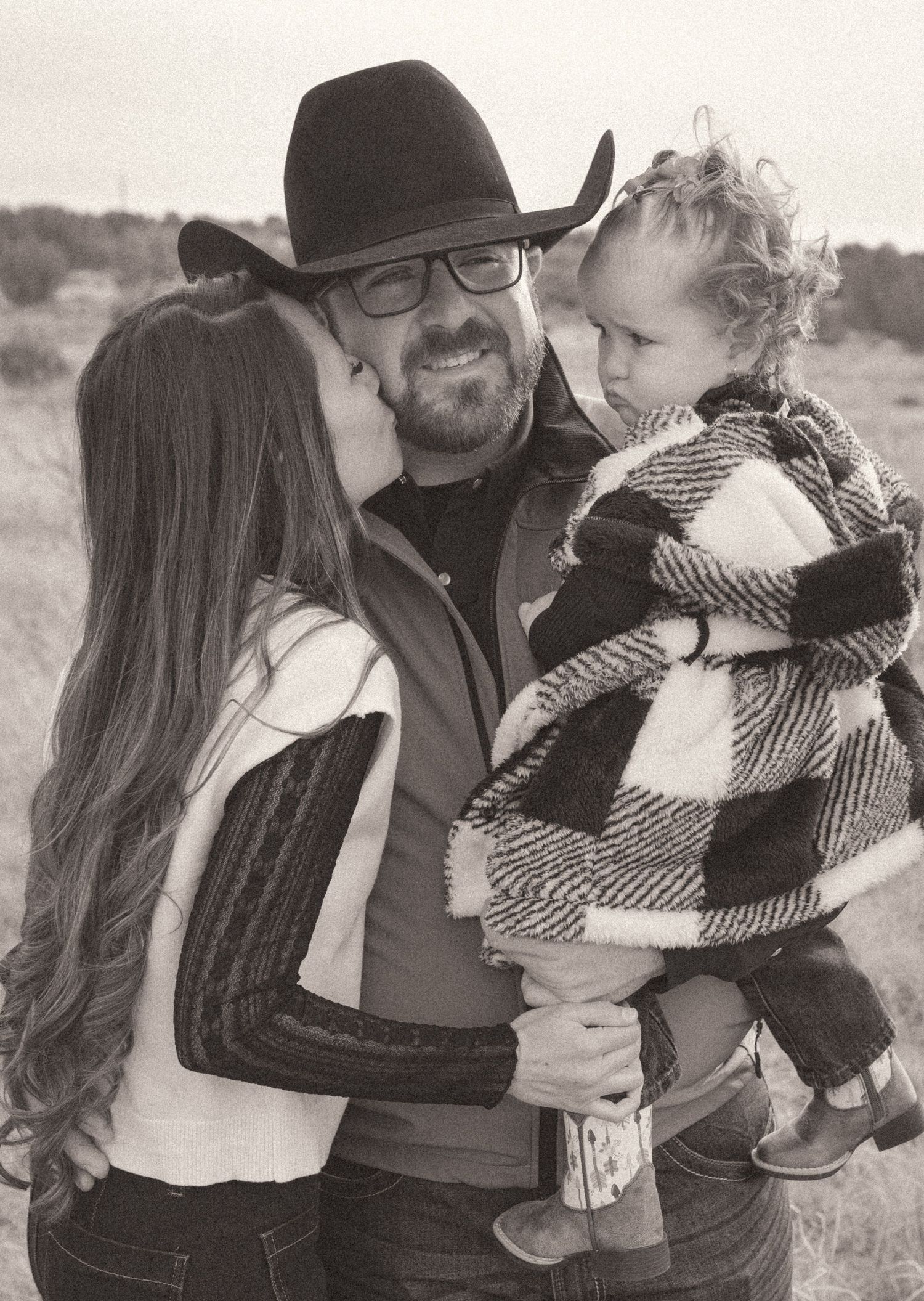 Black and white photo of couple with toddler, woman kissing man in cowboy hat.