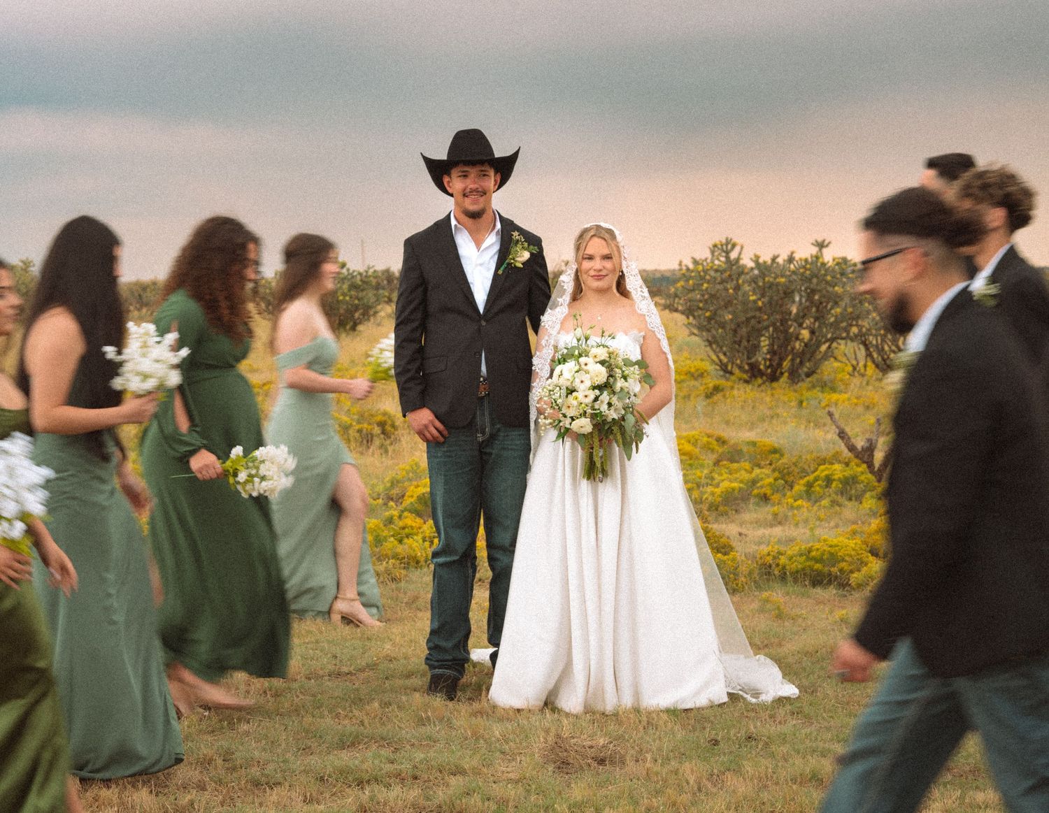 Bride and groom in cowboy hat at outdoor rustic wedding with green-dressed bridesmaids at sunset.