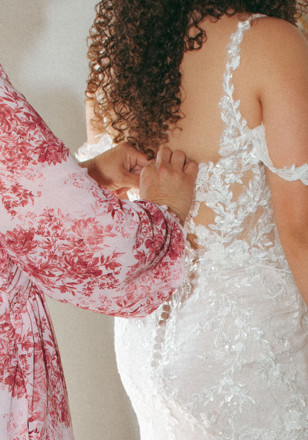 A woman in a floral dress fastens the lace back of a bridal gown before the wedding ceremony.