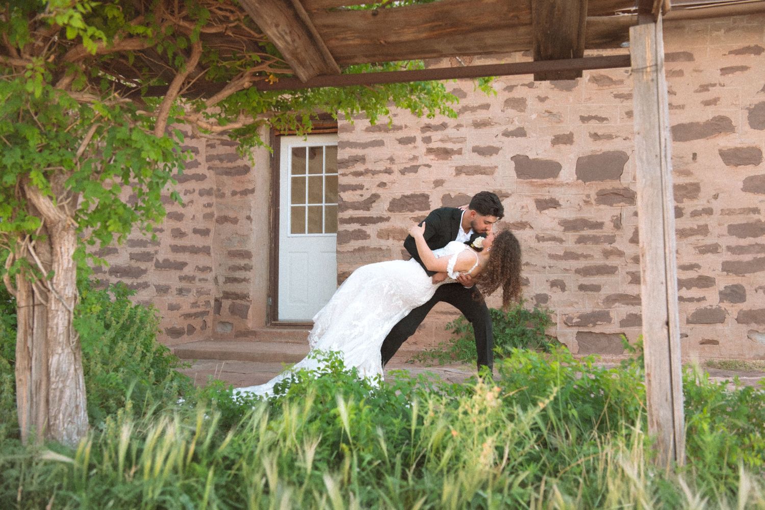 Couple sharing a romantic dip pose outside a rustic stone building surrounded by green foliage.
