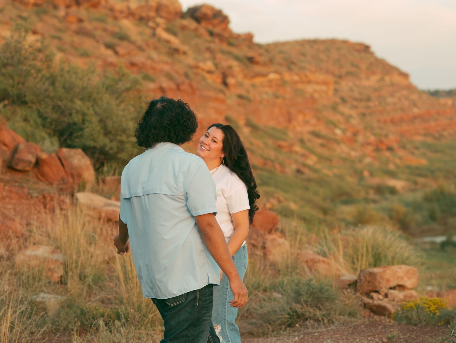 Smiling couple holding hands near red rocky hills at sunset in a candid outdoor portrait.