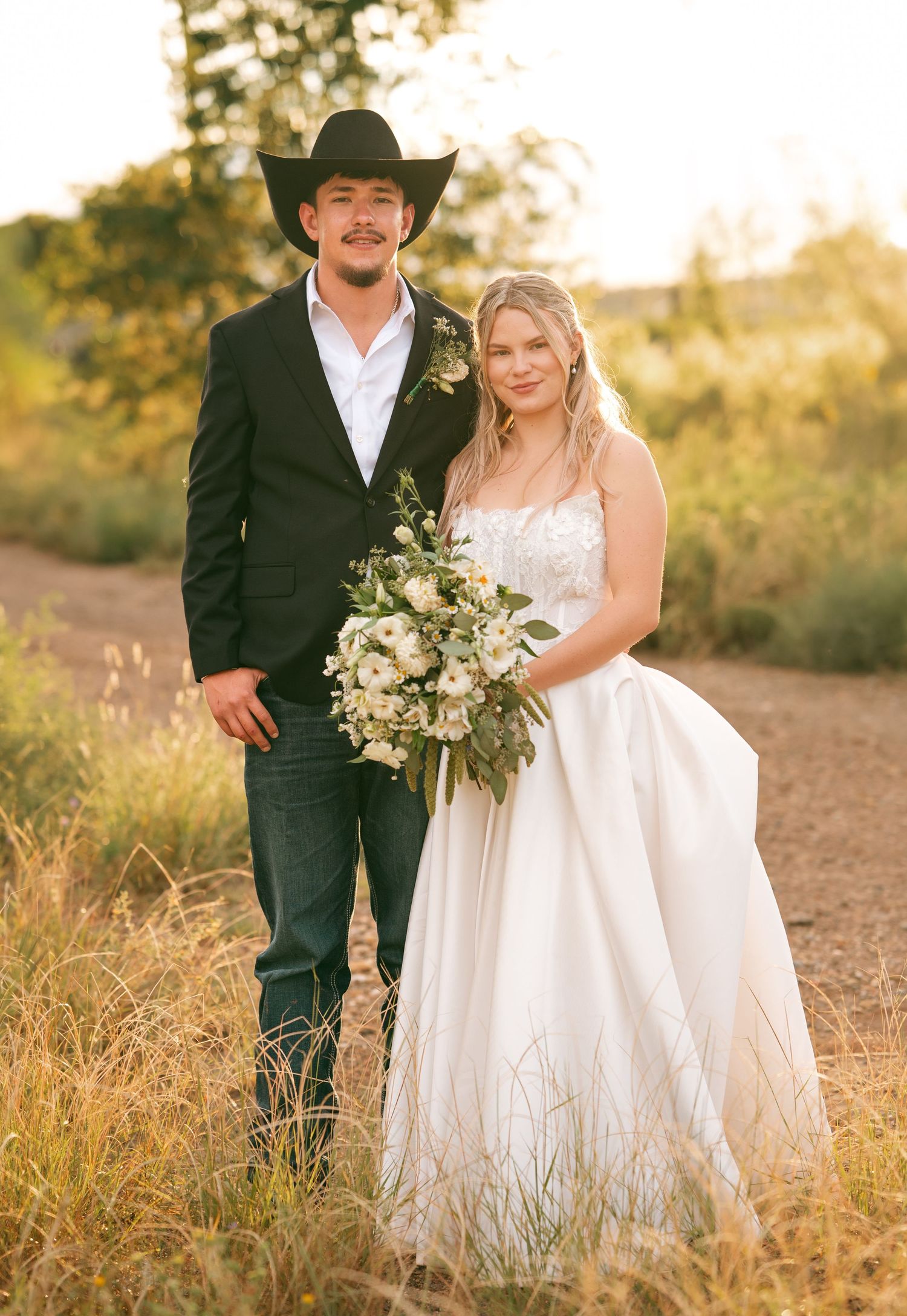 Bride and groom posing in golden hour light, groom in cowboy hat, bride holding white bouquet.