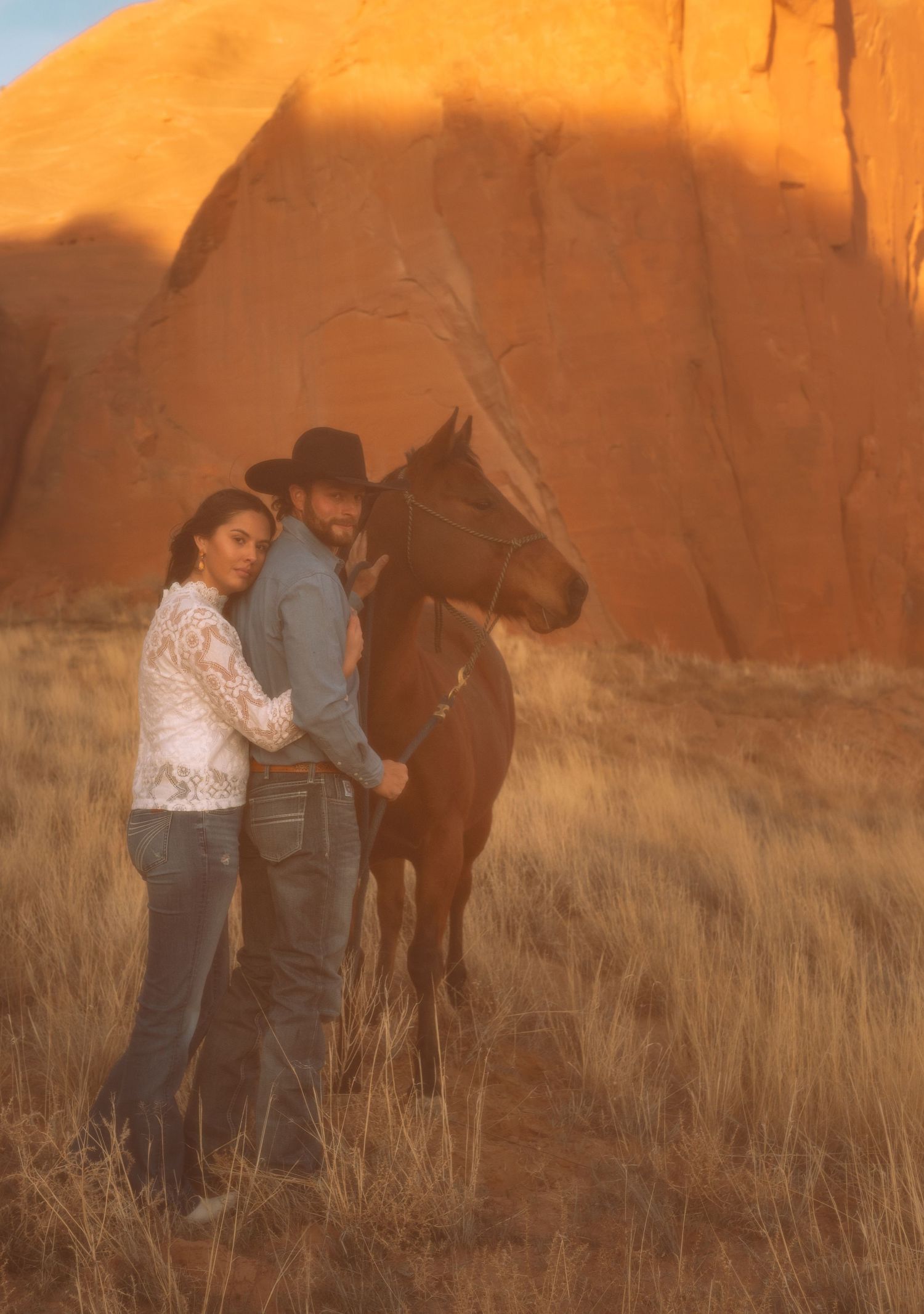 Couple standing with a horse against glowing red rock formations at golden hour in the desert.