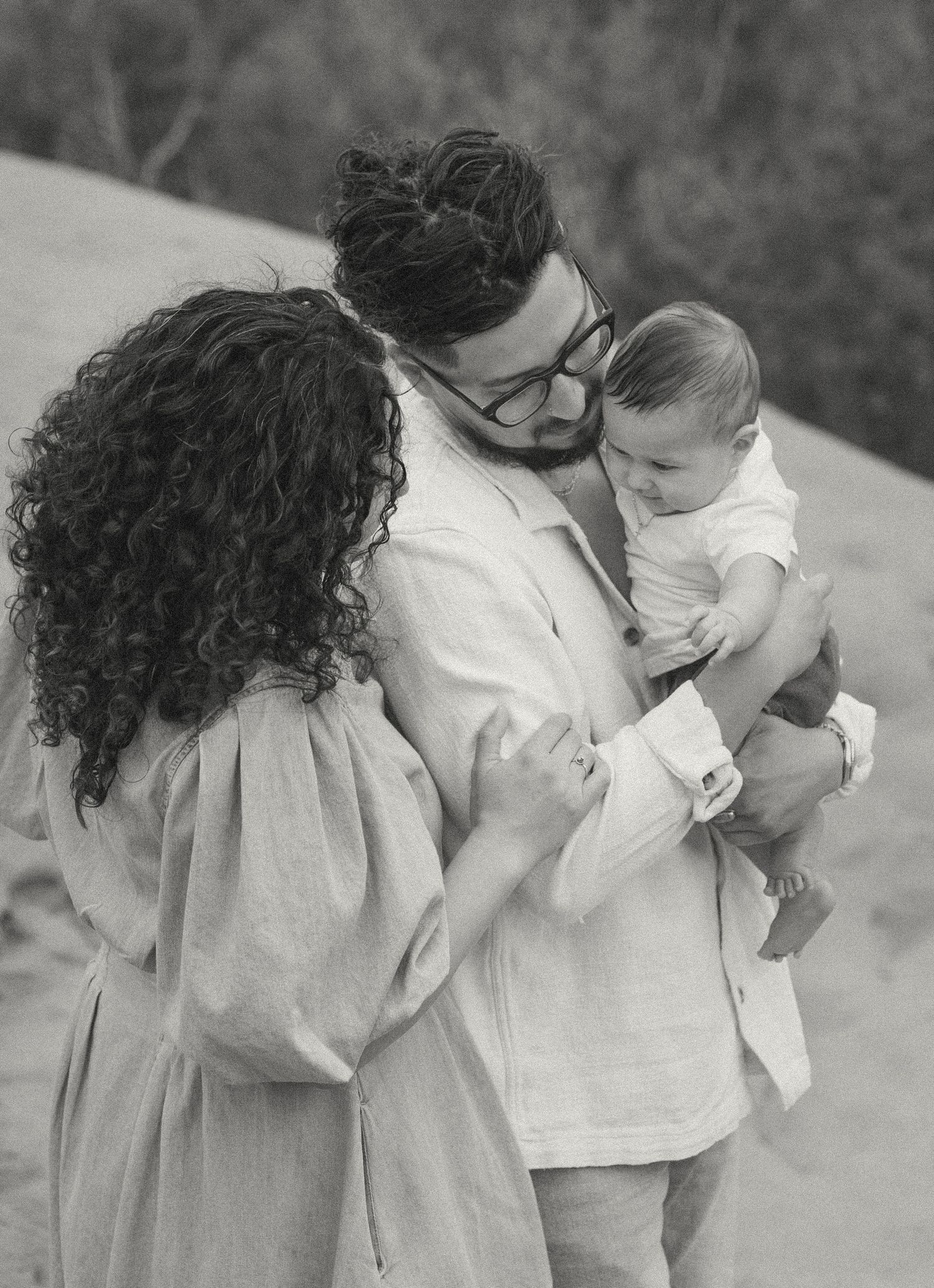 Black and white photo of couple sharing tender moment with baby in arms outdoors in natural setting.
