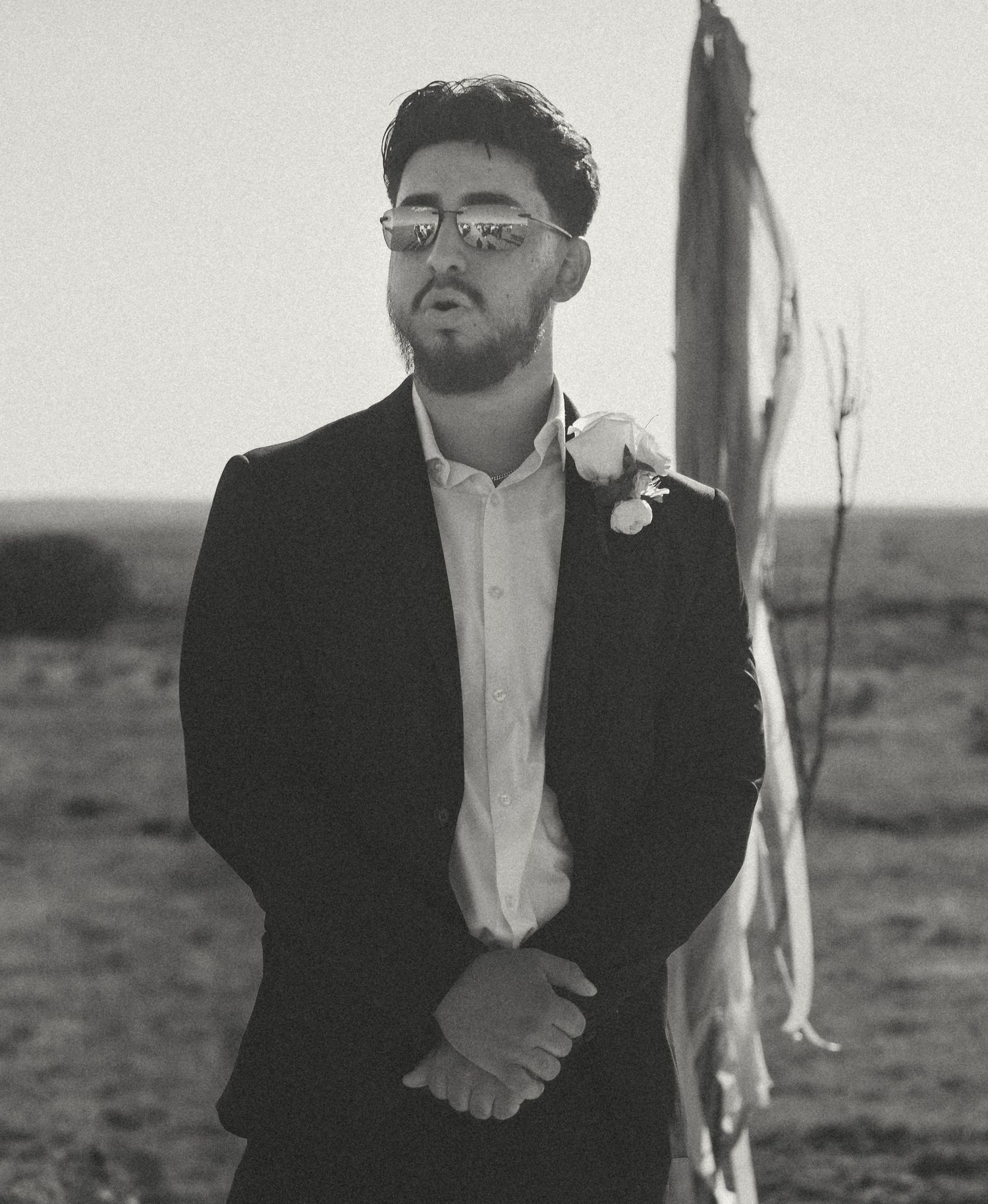 Groom in black suit and sunglasses stands solemnly outdoors before the wedding ceremony.