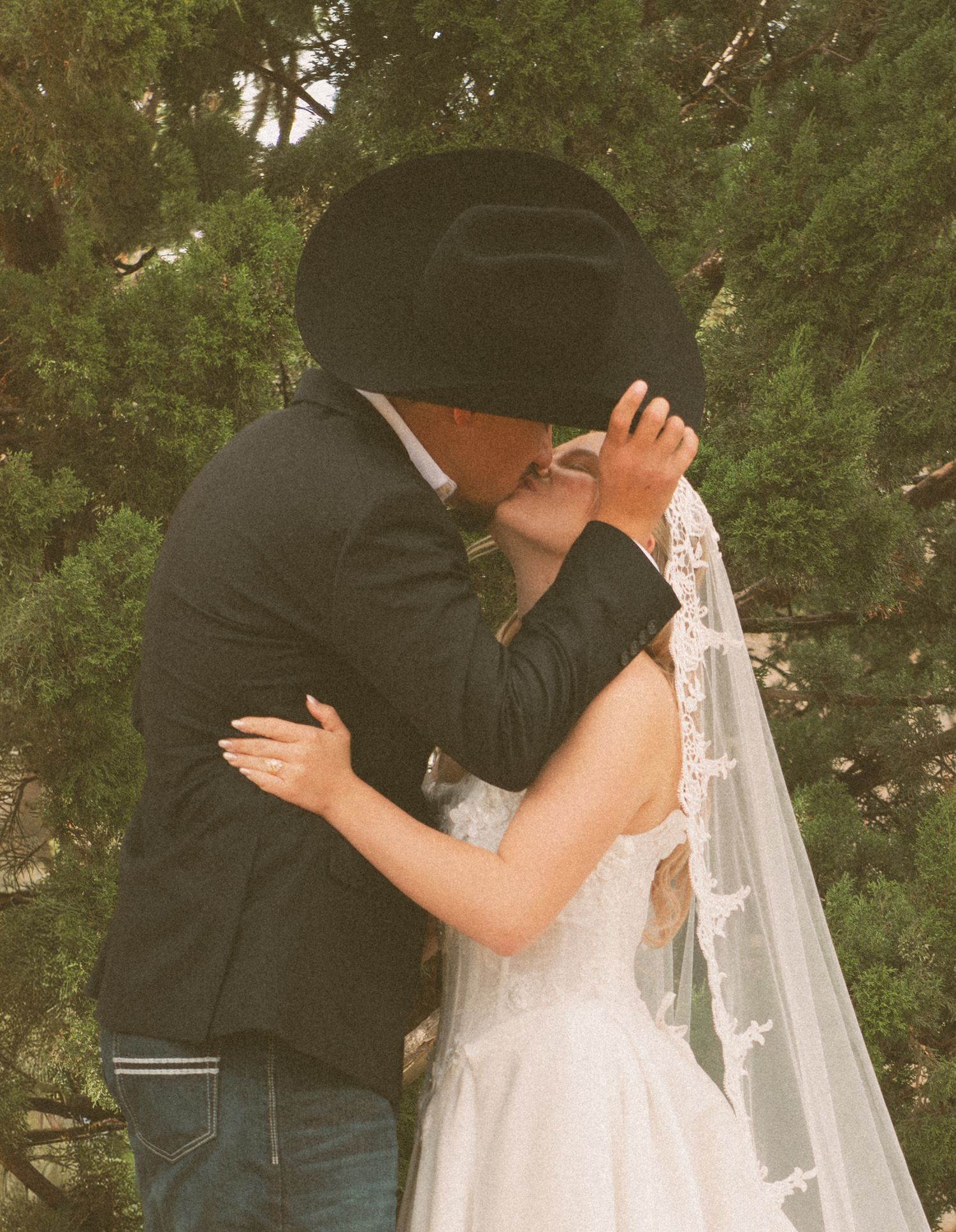 Groom in black cowboy hat and suit sharing first kiss with bride in lace wedding dress outdoors.