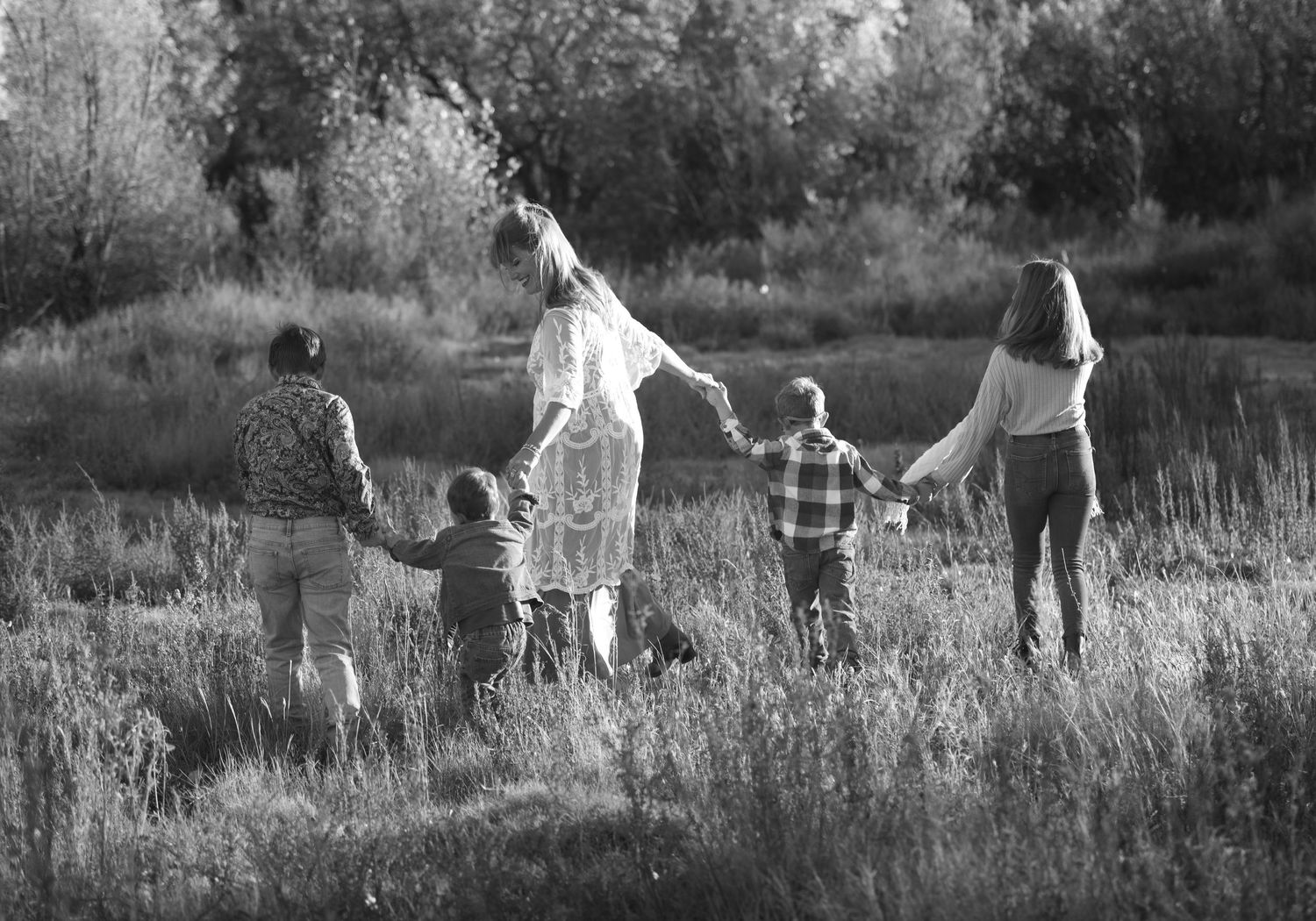 Black and white photo of a family of six holding hands walking through a lush meadow from behind.
