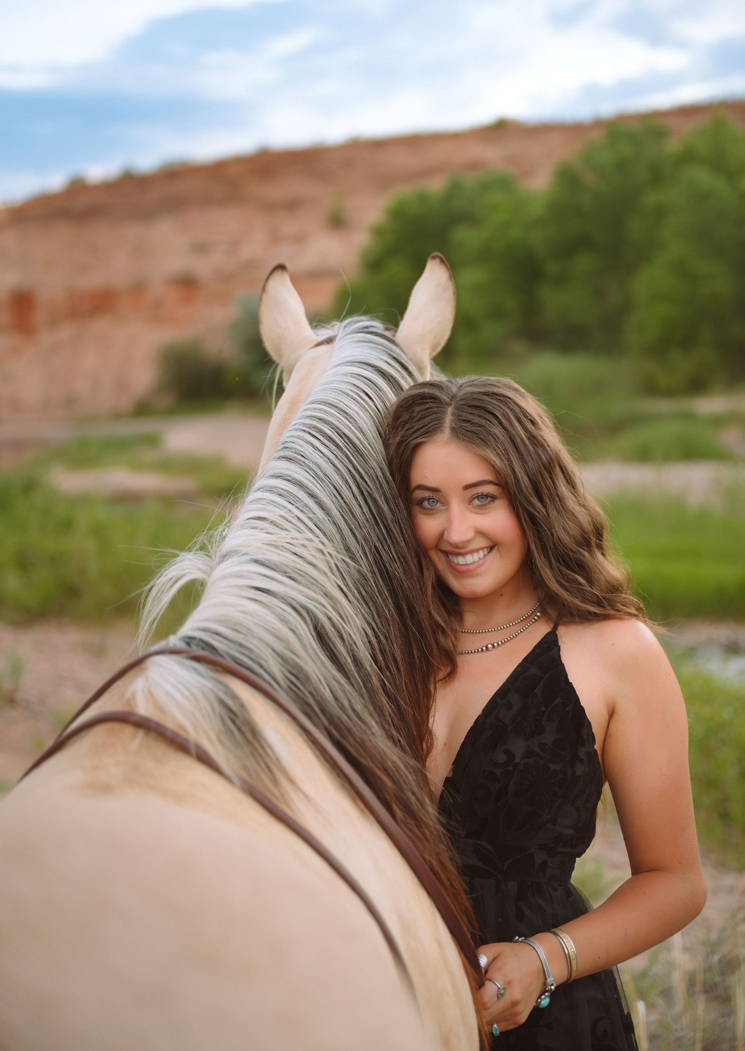 Smiling young woman in black dress posing with a gray horse in a scenic desert landscape with red rock formations.