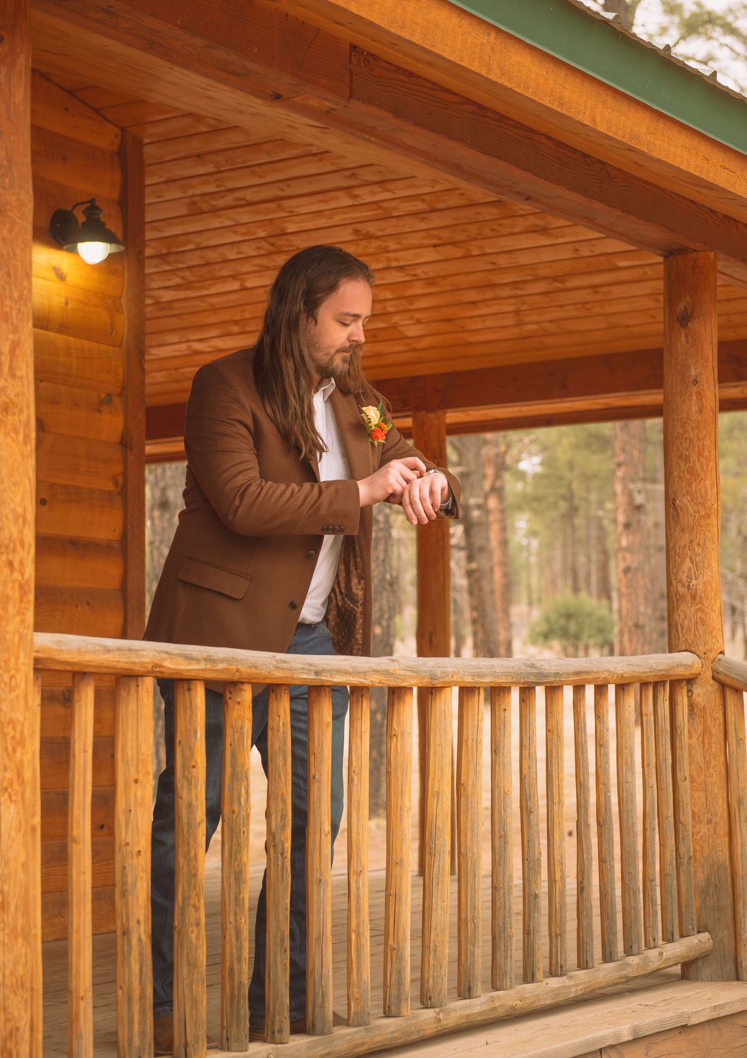 Groom in brown blazer adjusting boutonniere on rustic log cabin porch before wedding ceremony.