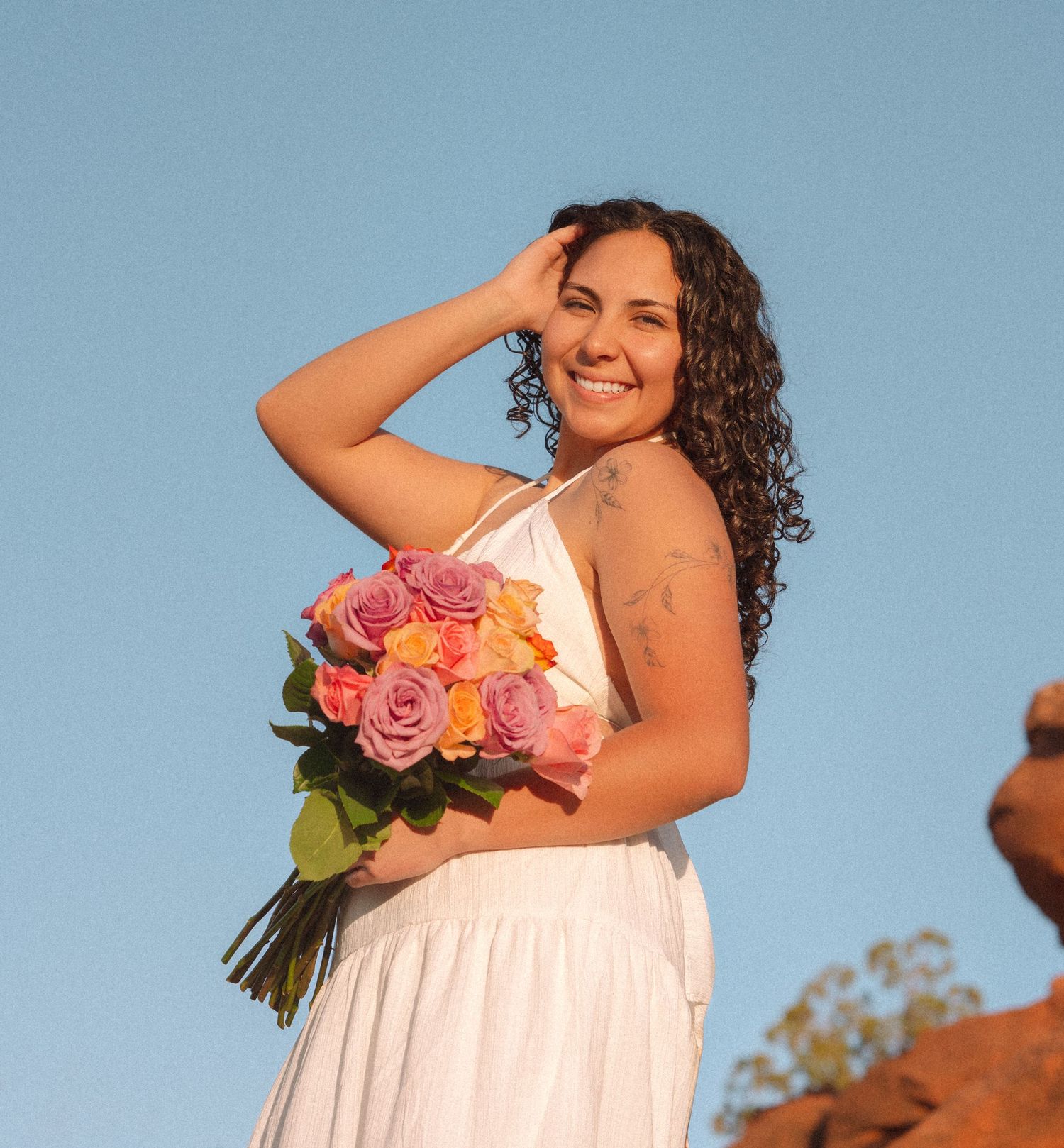 Smiling woman with curly hair holding colorful roses bouquet in white dress outdoors.