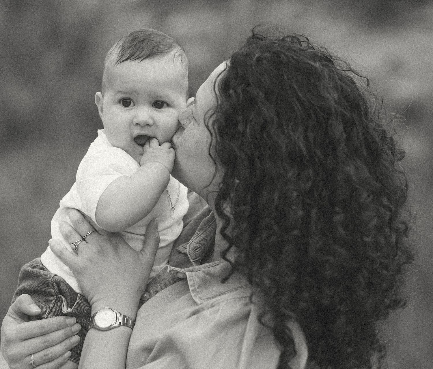 A curly-haired woman lovingly kisses a wide-eyed baby in a tender black and white portrait.