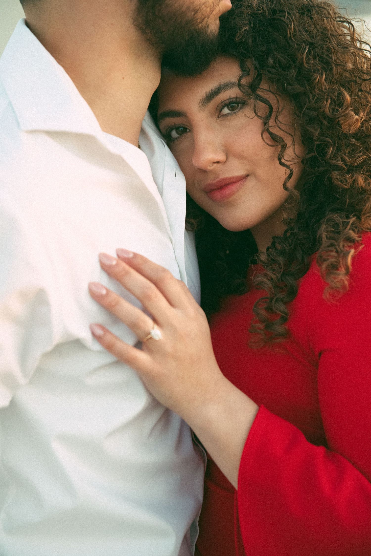 Woman with curly hair in red dress leaning against man in white shirt showing engagement ring.